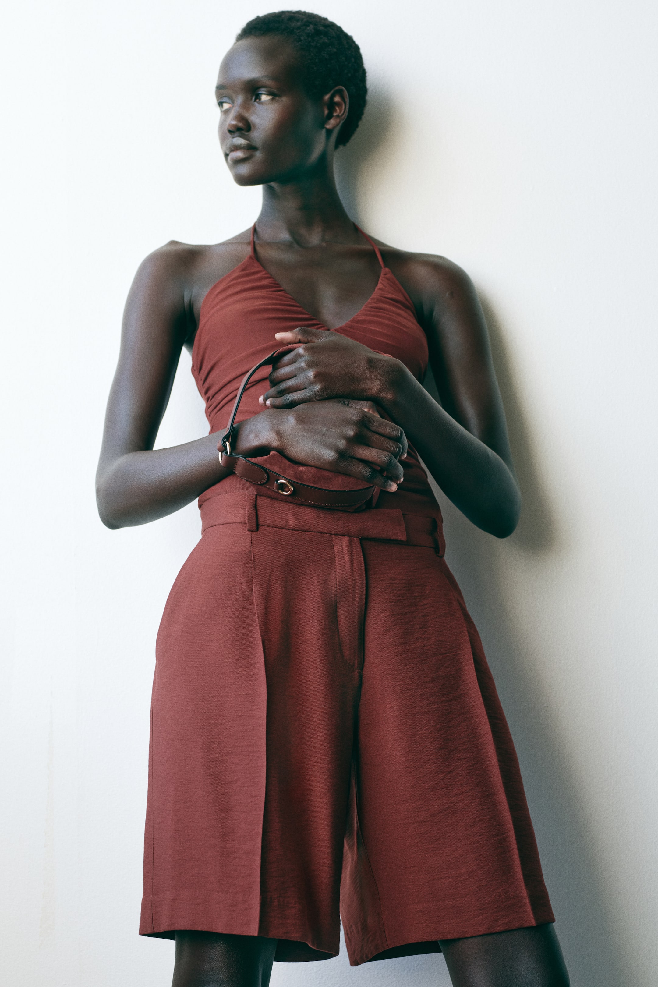 View larger image: A rust-colored halter neck top features a ruched bust and thin straps, paired with matching high-waisted, wide-leg shorts with front pleats. A small dark brown clutch with gold-toned hardware is held at the waist.