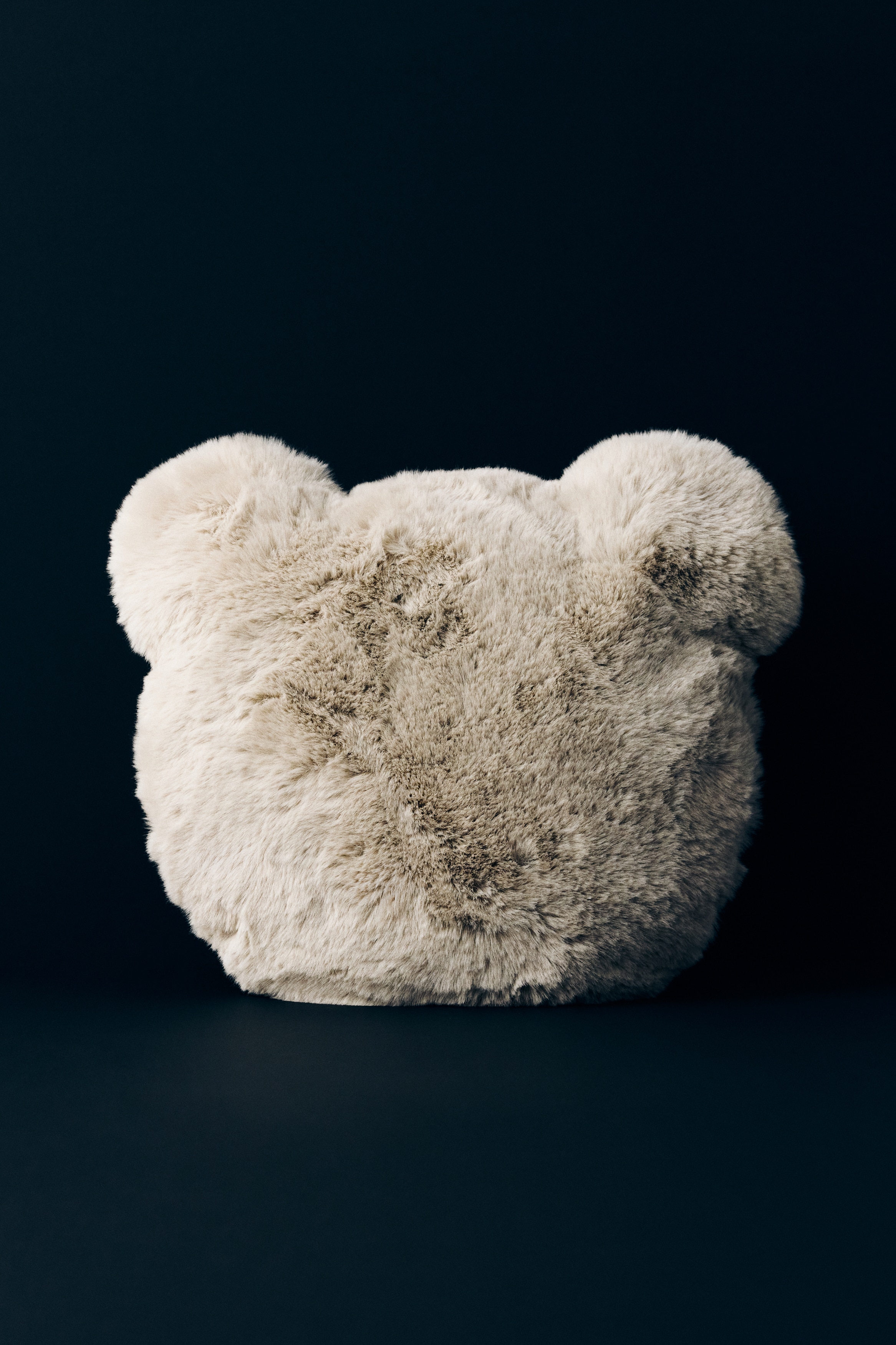 Un cuscino a forma di orso in un morbido tessuto effetto pelliccia beige chiaro, soffice e peloso, è rivolto frontalmente, mettendo in risalto la sua texture soffice. Il cuscino presenta una forma arrotondata con due piccole orecchie morbide nella parte superiore.