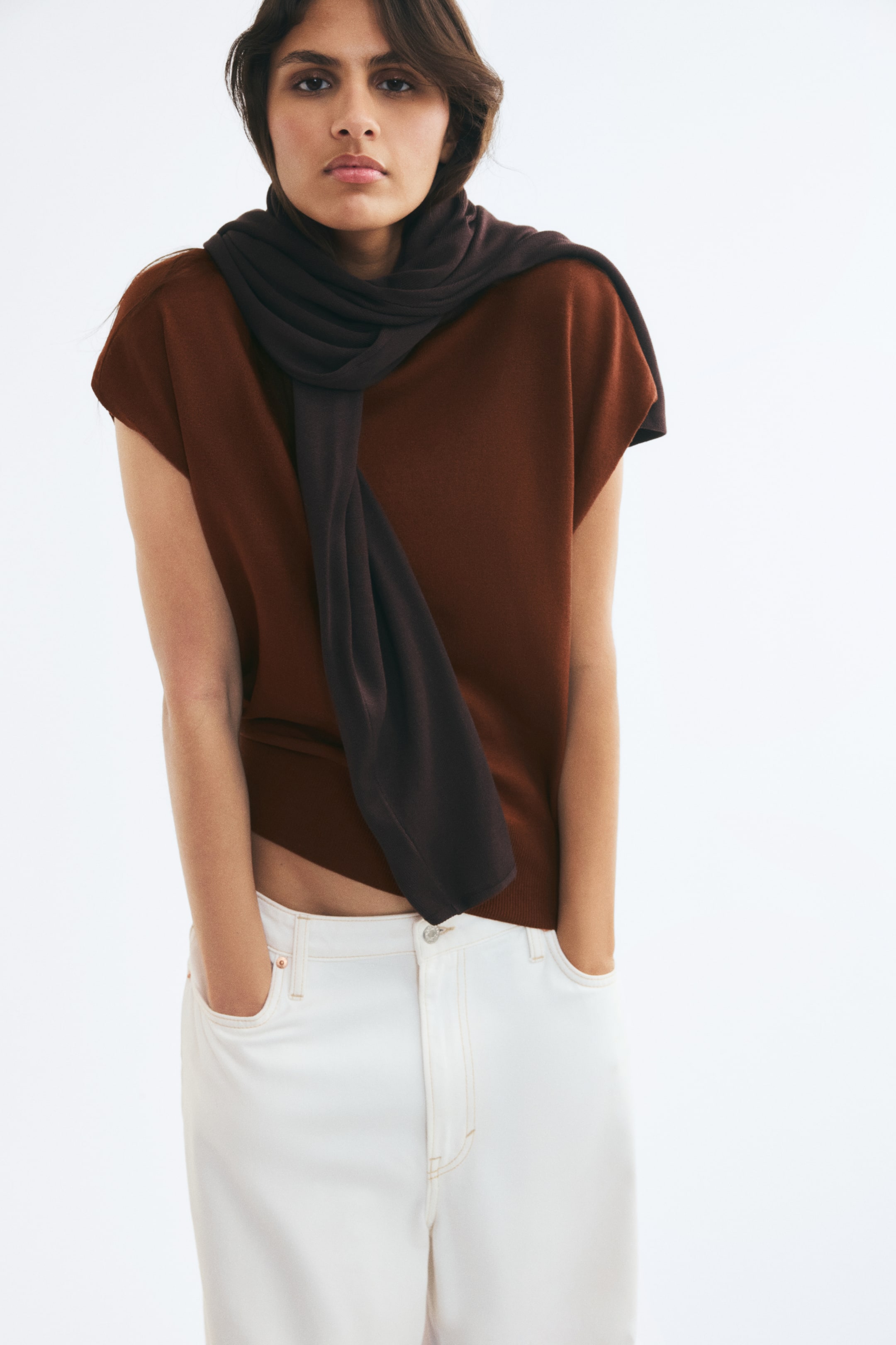 View larger image: A brown, fine-knit short vest with a high neck and ribbed hem is worn by the model. The vest is styled with a dark brown knit scarf draped around the neck and paired with white trousers.