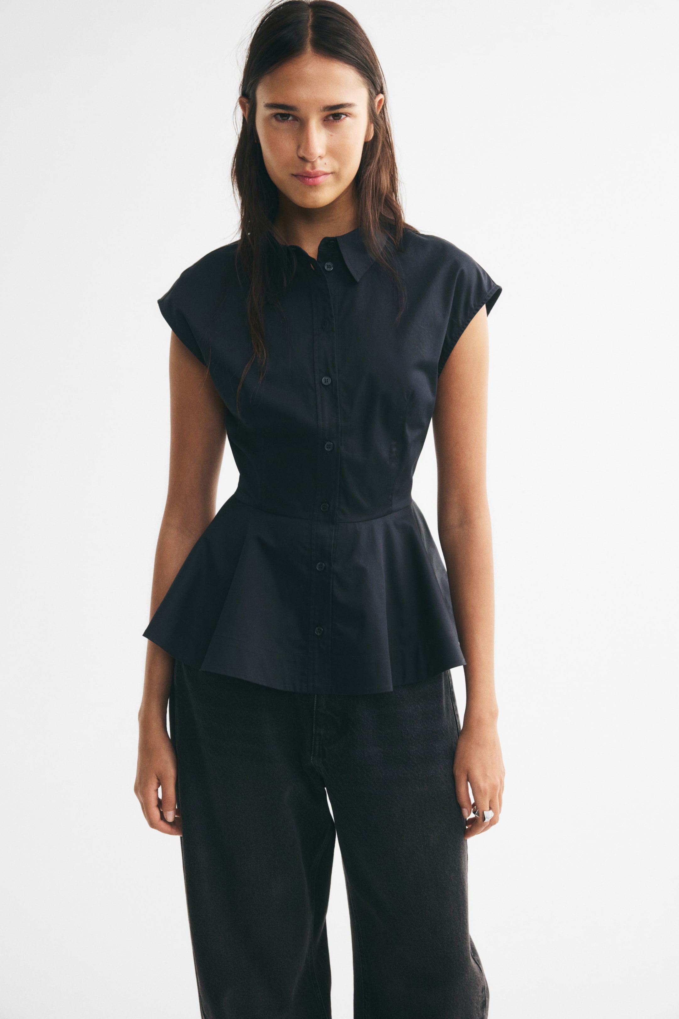 View larger image: A black sleeveless cotton poplin top featuring a collar, button-down front, waist darts, and a flared peplum is being worn by the model. The top is styled with dark gray jeans.