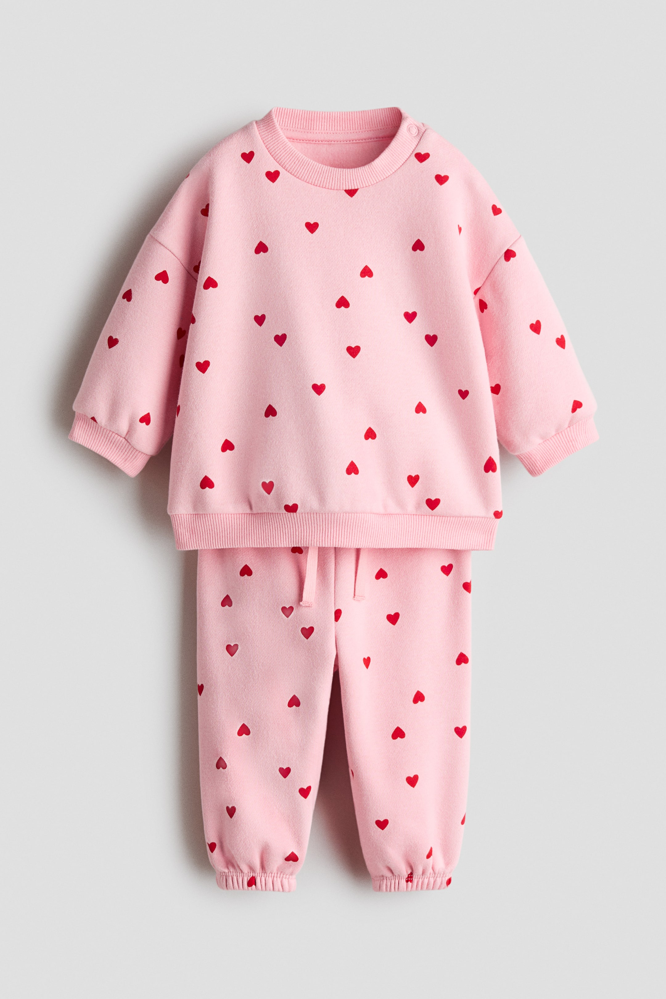 View larger image: A pink tracksuit is displaying a sweatshirt and matching sweatpants, both patterned with scattered red hearts. The sweatshirt is featuring a ribbed crew neck, ribbed cuffs, and a ribbed hem, with snap buttons on one shoulder. The sweatpants are showcasing an elasticated drawstring waist and ribbed ankle cuffs.