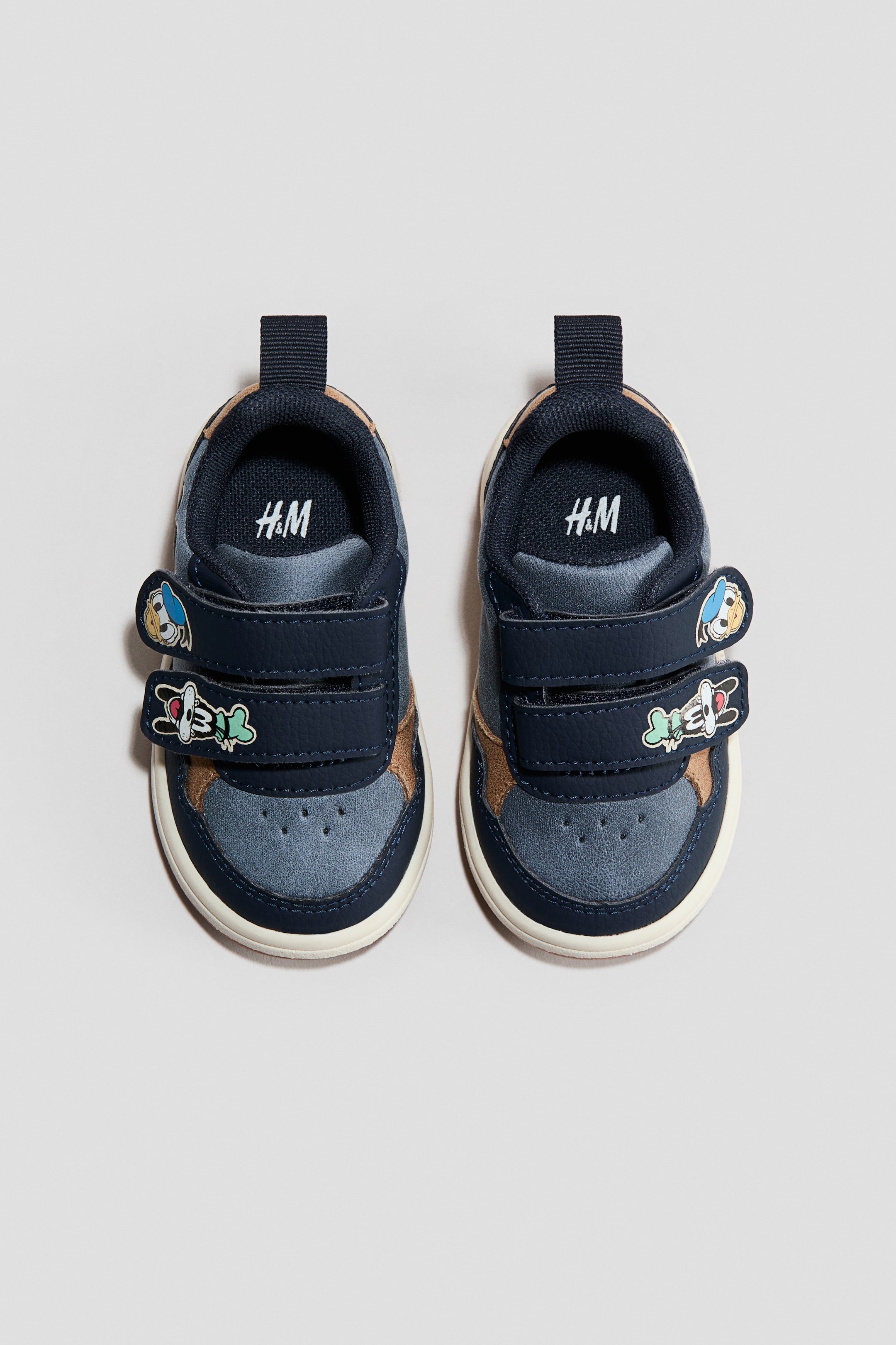 Ampliar la imagen: Un par de tenis para bebé en color azul marino se presenta de FRENTE, exhibiendo paneles de cuero sintético y similares al ante en azul marino, azul claro y beige. Cada zapato cuenta con dos correas de gancho y bucle, adornadas con aplicaciones de Donald Duck y Goofy, y descansa sobre una suela de goma de color crema.