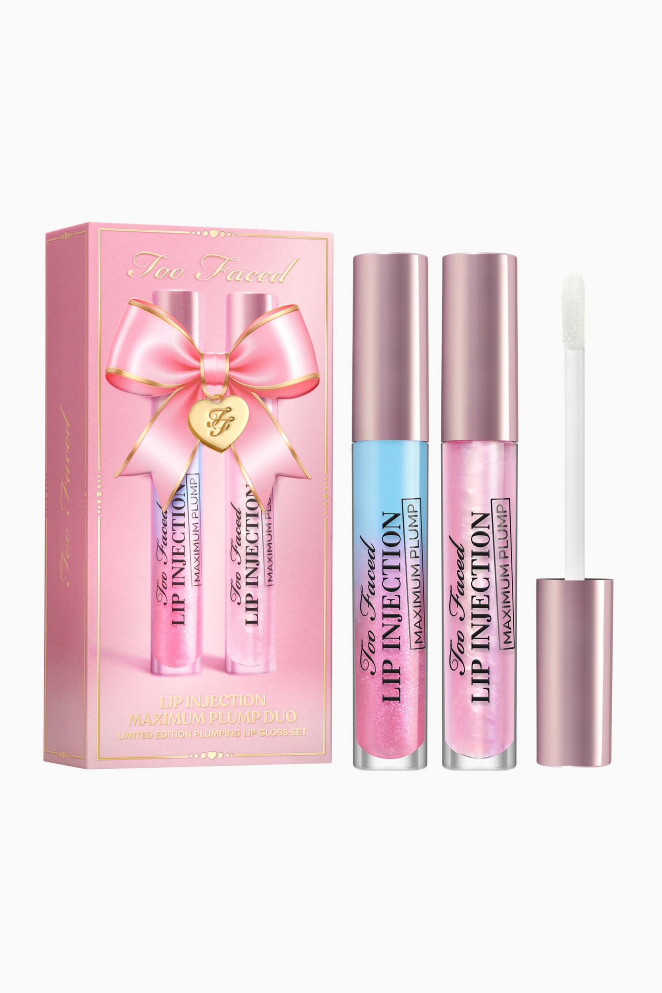 Näytä suurempi kuva: Too Faced Lip Injection Maximum Plump Duo Set -lahjapakkaus sisältää vaaleanpunaisen suorakulmaisen lahjarasian, jota koristaa vaaleanpunainen rusetti, sekä kaksi pystyasennossa olevaa huulikiiltoputkiloa ja avoimen applikaattoripuikon. Toinen kirkas putkilo sisältää sinisestä pinkkiin hohtavaa nesteen sävyä, kun taas toisessa on vaaleanpunainen, kimalteleva neste, ja molempia viimeistelevät metallinhohtoiset vaaleanpunaiset korkit. Valkoinen doe-foot-applikaattori seisoo oikealla pystyssä, korostaen sen muotoa ja designia.