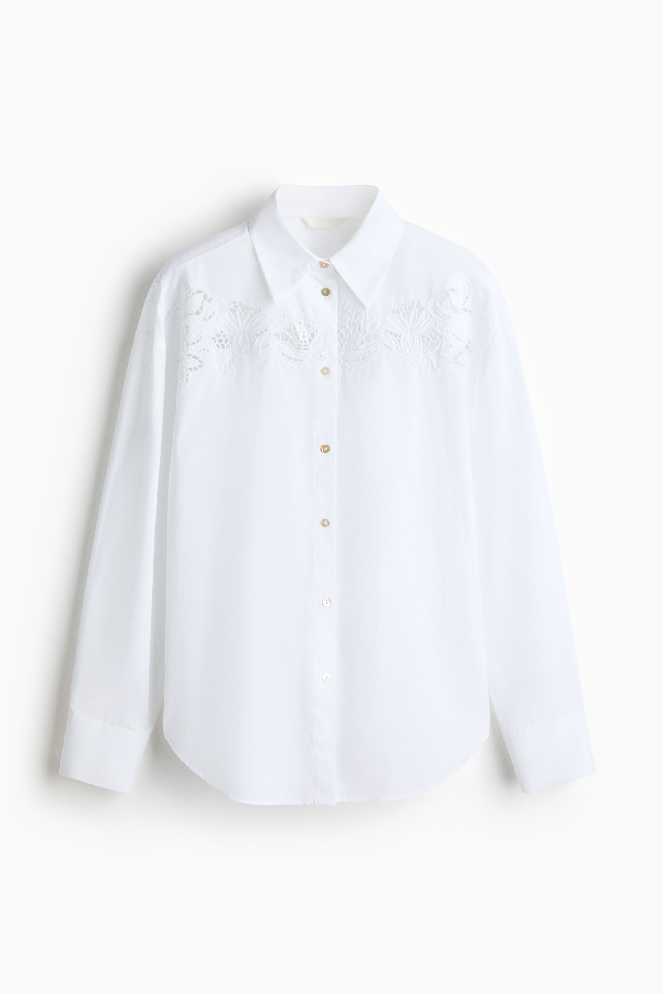 Ampliar la imagen: Se presenta una camisa blanca de manga larga desde el frente, con cuello en punta y una tapeta de botones con botones de color claro. Un bordado intrincado de encaje recortado adorna el pecho y los hombros, y la camisa termina con un dobladillo redondeado.