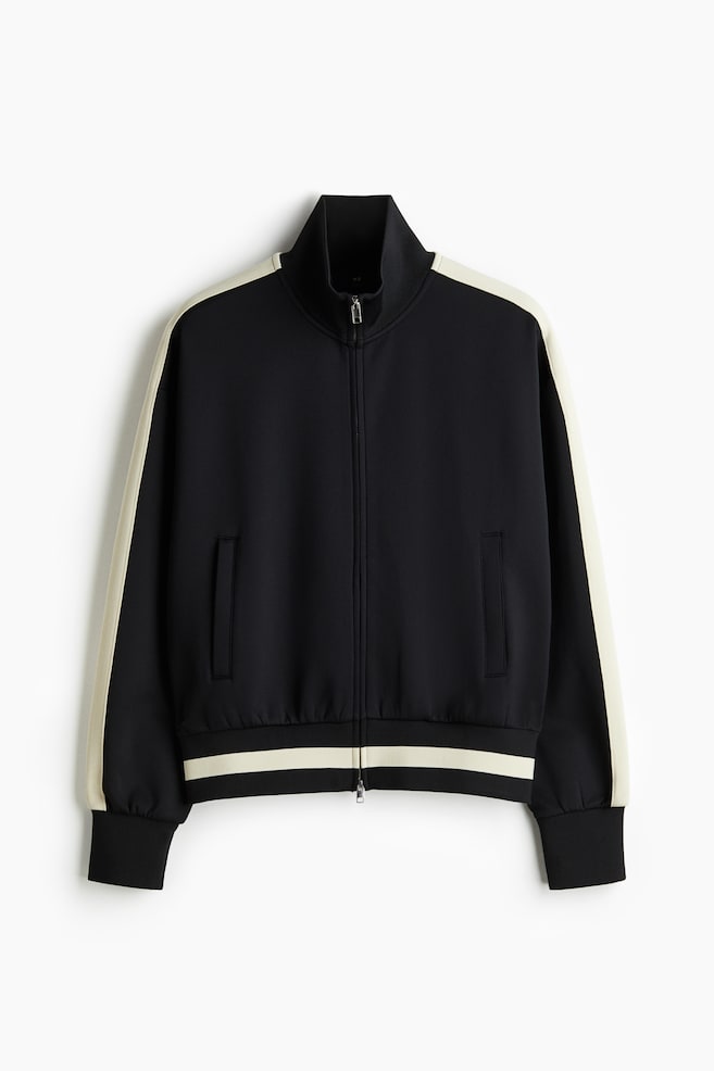 Eine schwarze Trainingsjacke wird von vorne gezeigt, mit einem Stehkragen, einem durchgehenden Reißverschluss mit silbernem Reißverschluss und zwei vertikalen Schlitztaschen. Off-weiße Streifen verzieren die Außenseiten der langen Ärmel und ein Doppelstreifen ist am gerippten Saum sichtbar, der mit den gerippten Bündchen koordiniert. - 1