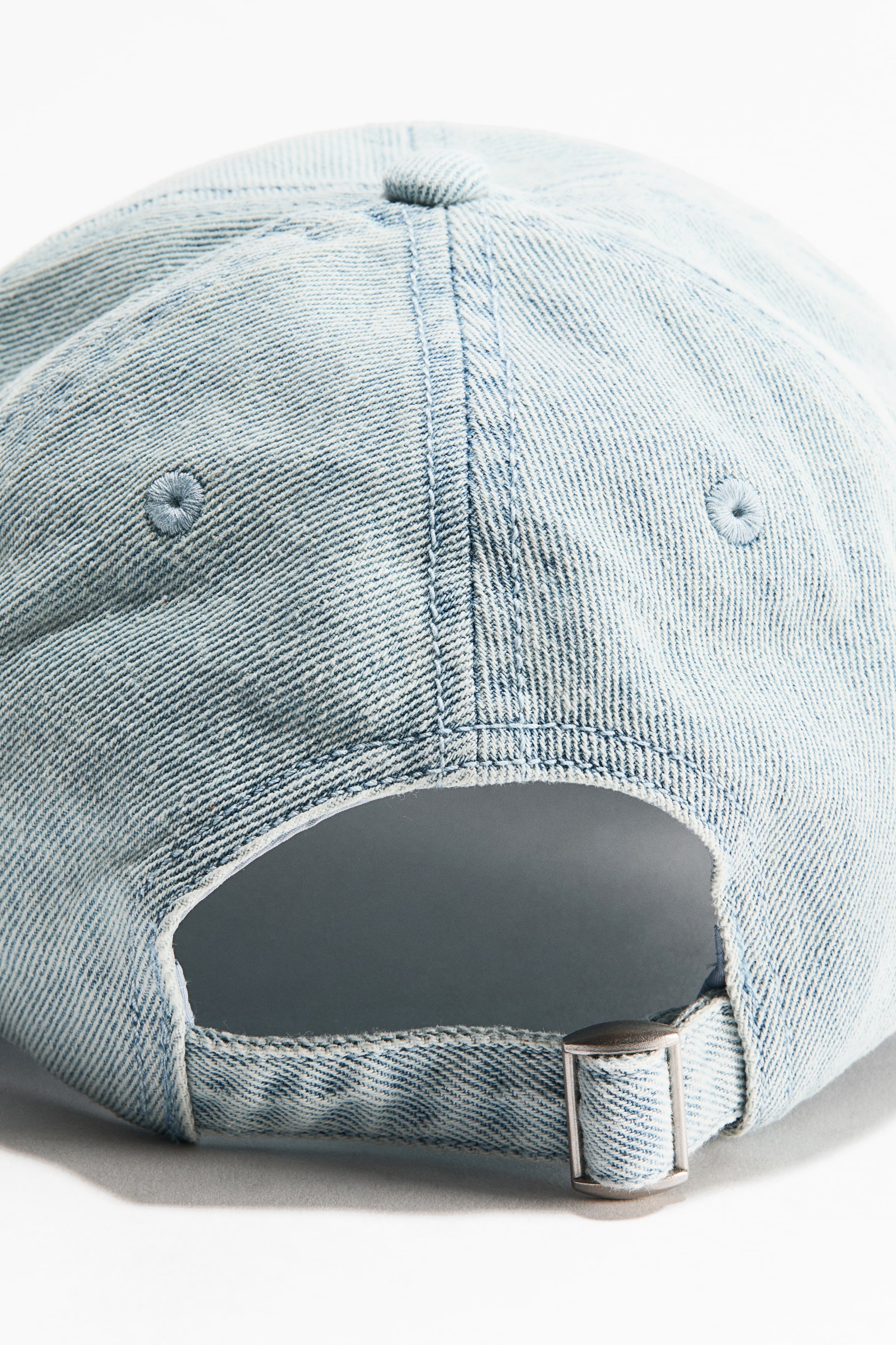 Vezi imaginea mai mare: O șapcă din bumbac twill de culoare albastru deschis este prezentată dintr-o vedere din spate, având cusături vizibile, orificii de ventilație pe panourile laterale și o bandă reglabilă din material textil cu o cataramă metalică de culoare argintie.