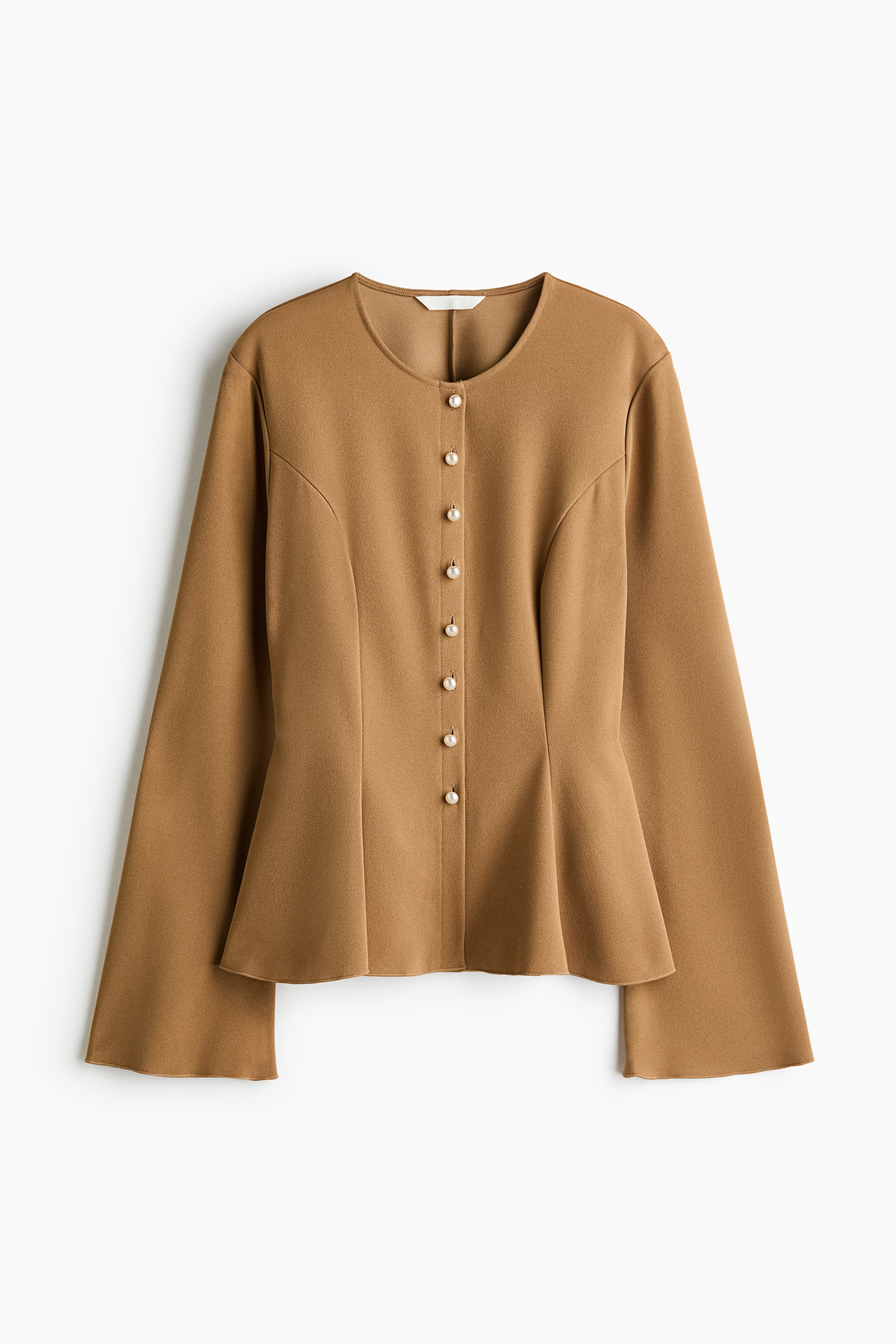 A camel-coloured jacket is worn by the model, featuring a collarless neckline, flared long sleeves, and a structured peplum hem. The jacket is styled with blue jeans, showcasing its tailored side profile.