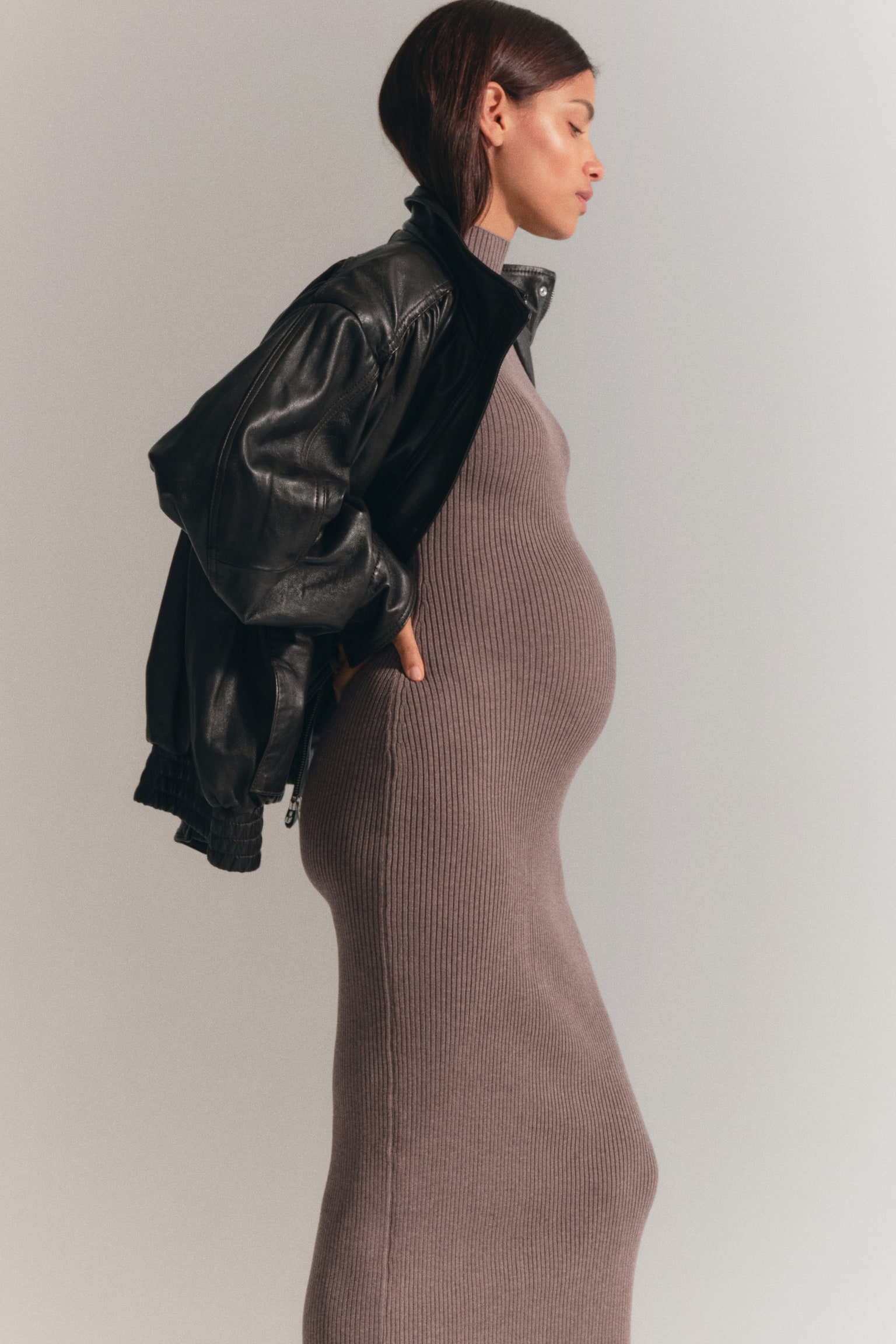 A long, fitted dress in taupe rib knit with a high neck is being worn by the model. A black leather jacket is layered over the shoulders of the dress. - 1