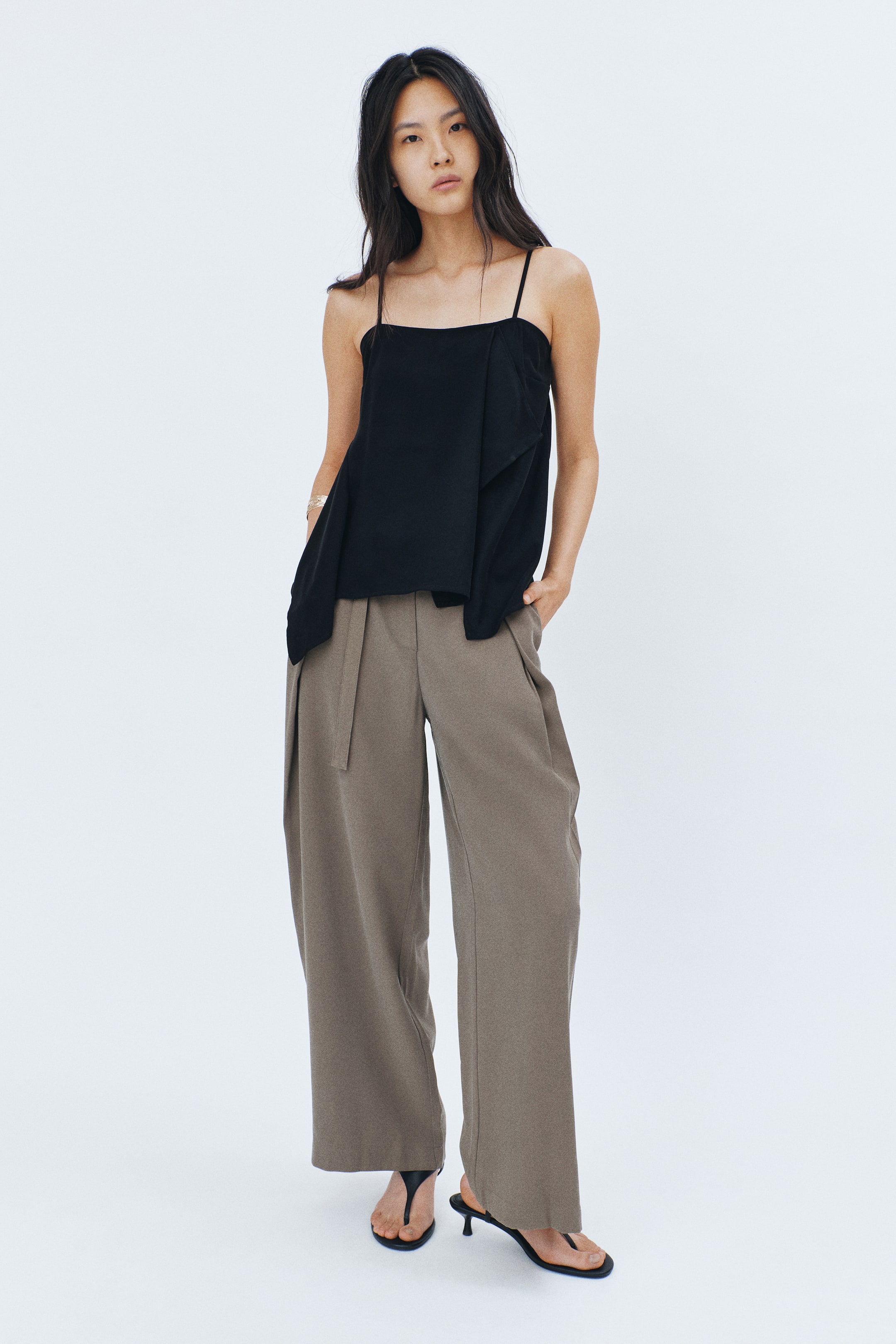 View larger image: A black camisole top with thin straps, an asymmetrical hem, and draped detailing is being worn. The model is also wearing brown pleated wide-leg pants with a waist tie, standing with her hands in the pockets.
