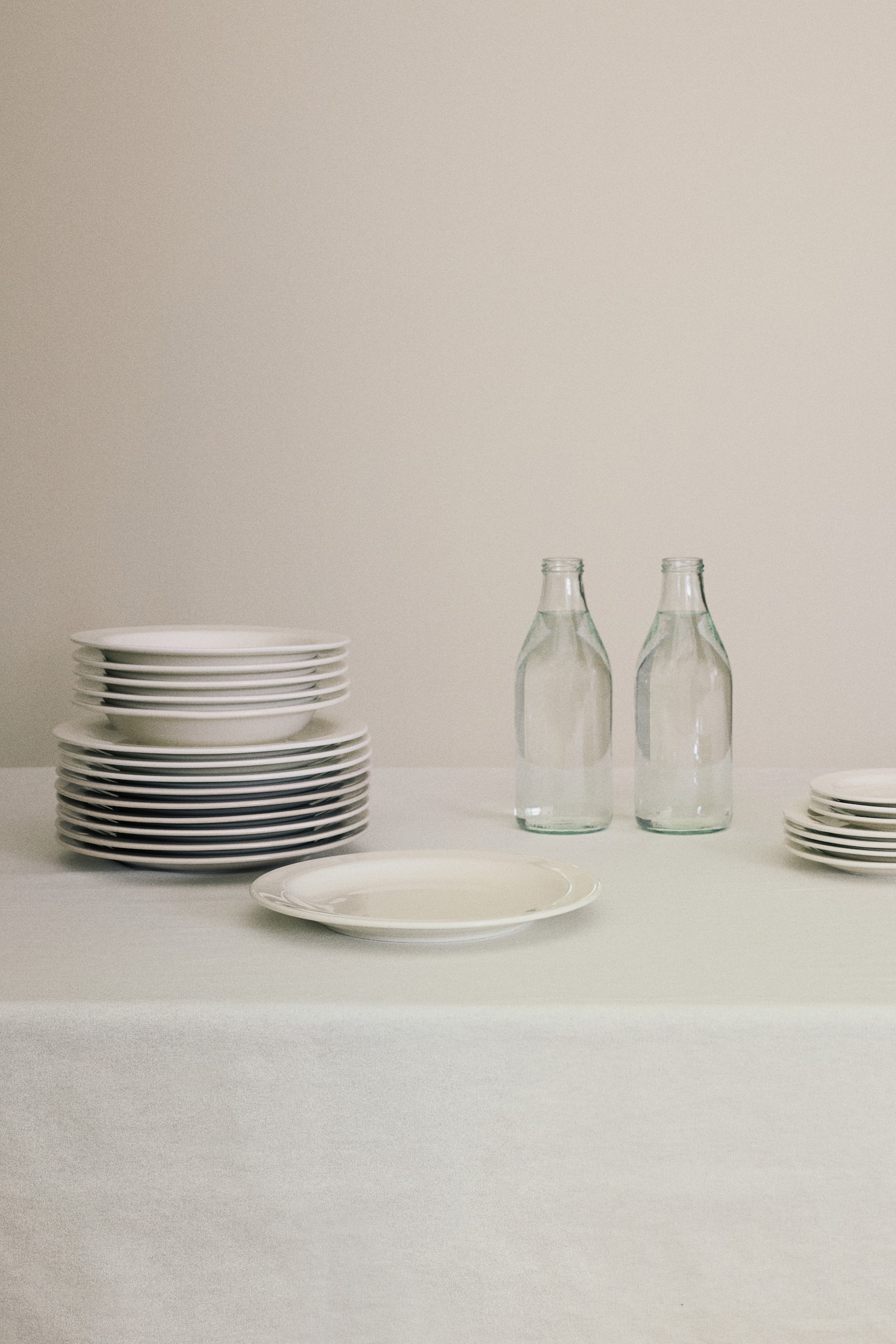 A single off-white porcelain dinner plate with a minimalist design and a classic rim lies flat in the foreground. To the left, a tall stack of the dinner plates supports a smaller stack of matching rimmed bowls, highlighting their clean, uniform structure. - 1