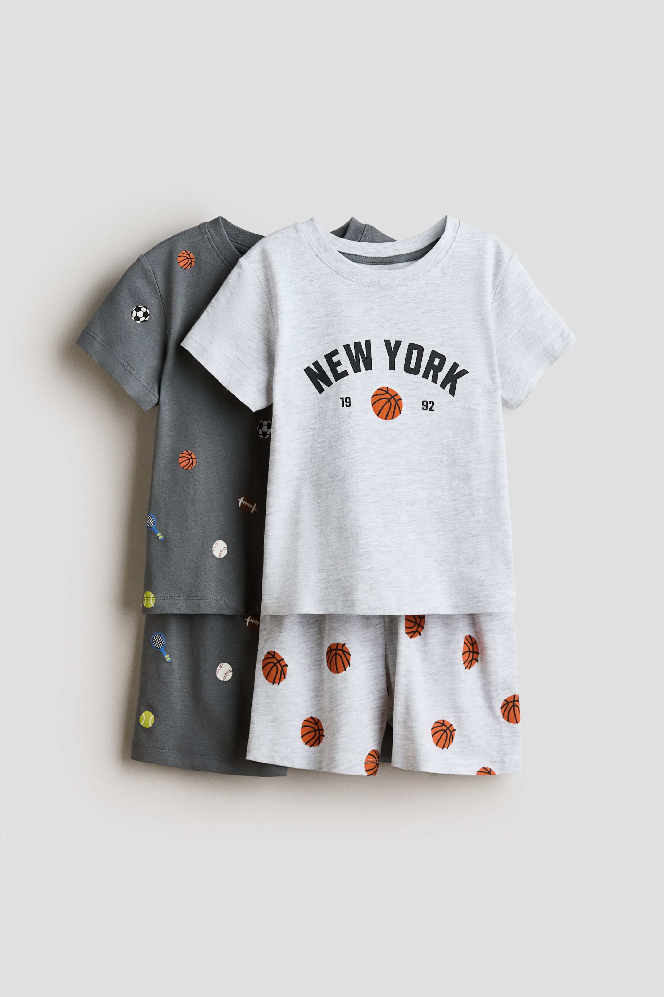 View larger image: Pyjamas with a T-shirt and matching shorts are displayed in two colourways. The light grey set features a T-shirt with a ribbed crew neckline and a 'NEW YORK' basketball graphic, along with shorts showcasing an all-over basketball print. The dark grey set, partially visible in the background, displays a T-shirt and shorts with an all-over print of various sports balls.