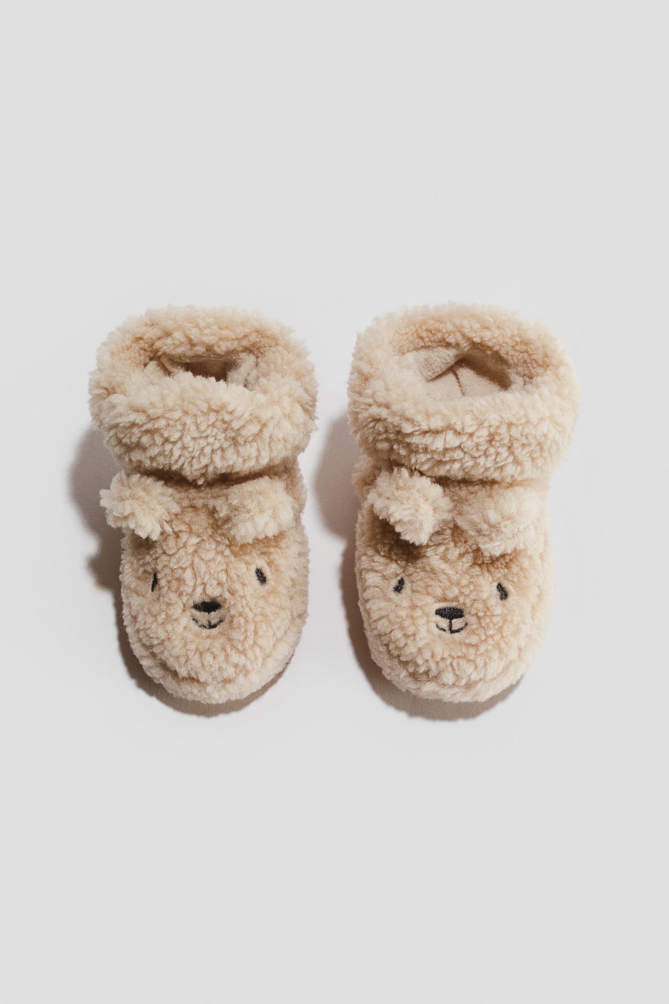 View larger image: A pair of tan sherpa baby booties stands upright, facing forward. Each bootie showcases an embroidered bear face with black eyes and a nose, complemented by small floppy ears attached to the sides.