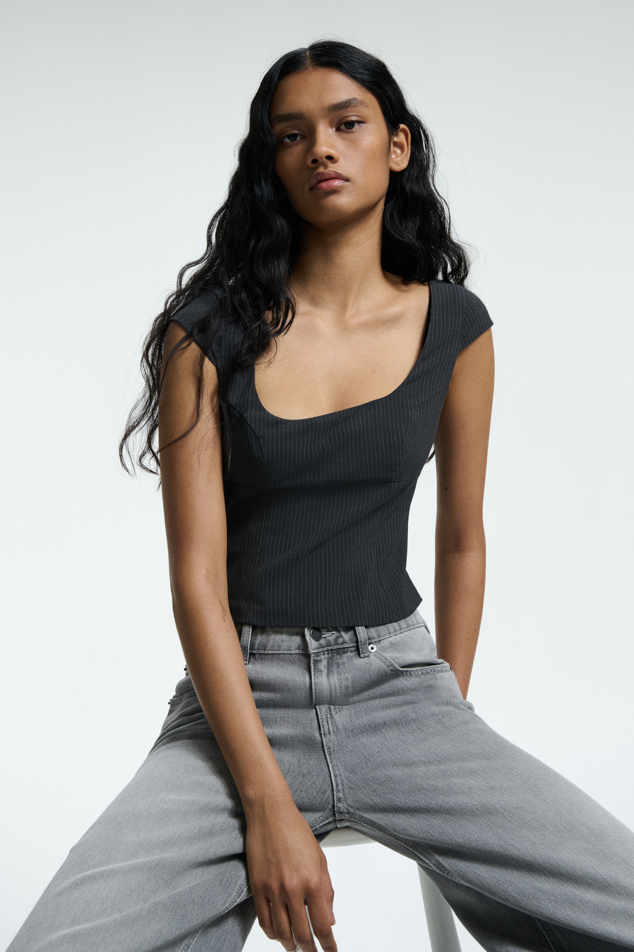 Ver imagen más grande: Se presenta una parte superior de color gris oscuro desde una vista frontal, con finas rayas verticales. Exhibe un escote redondeado estructurado y mangas cortas tipo capa, con una silueta ajustada que termina en la cintura.
