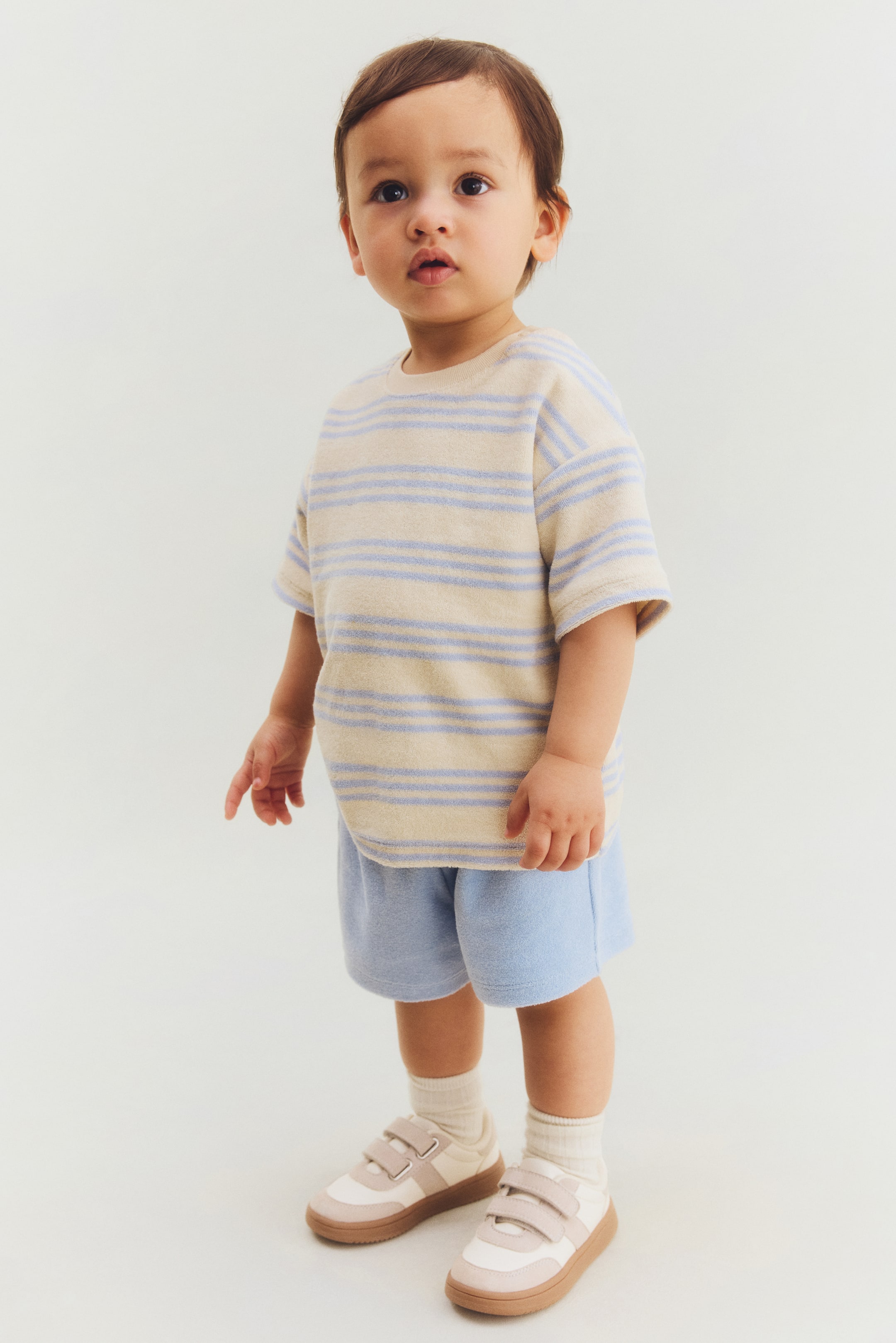 Nagyobb kép megtekintése: Egy póló és rövidnadrág szett kerül bemutatásra elölről. A póló krém- és világoskék vízszintes csíkokkal, bordázott kerek nyakkivágással és ejtett vállakkal rendelkezik. A világoskék rövidnadrág rugalmas derékpánttal van ellátva, és mindkét darab puha frottír anyagból készült.