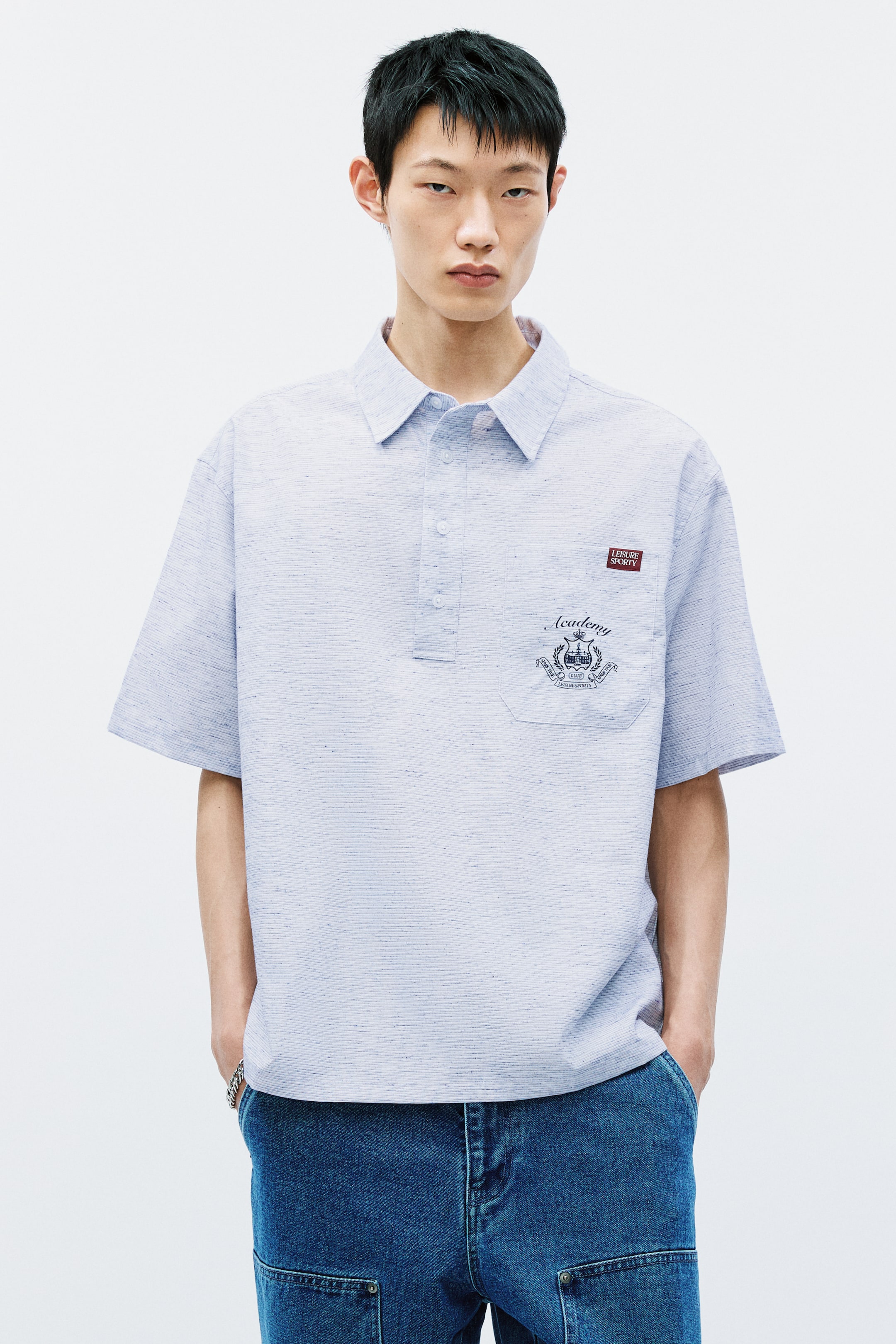 Xem ảnh lớn hơn: Một chiếc áo polo ngắn tay màu xanh nhạt với họa tiết heathered đang được giới thiệu, mặt trước, có cổ tiêu chuẩn và hàng khuy ba nút. Túi vá ngực trái hiển thị thiết kế huy hiệu thêu, với nhãn "LEISURE SPORTY" xuất hiện phía trên.