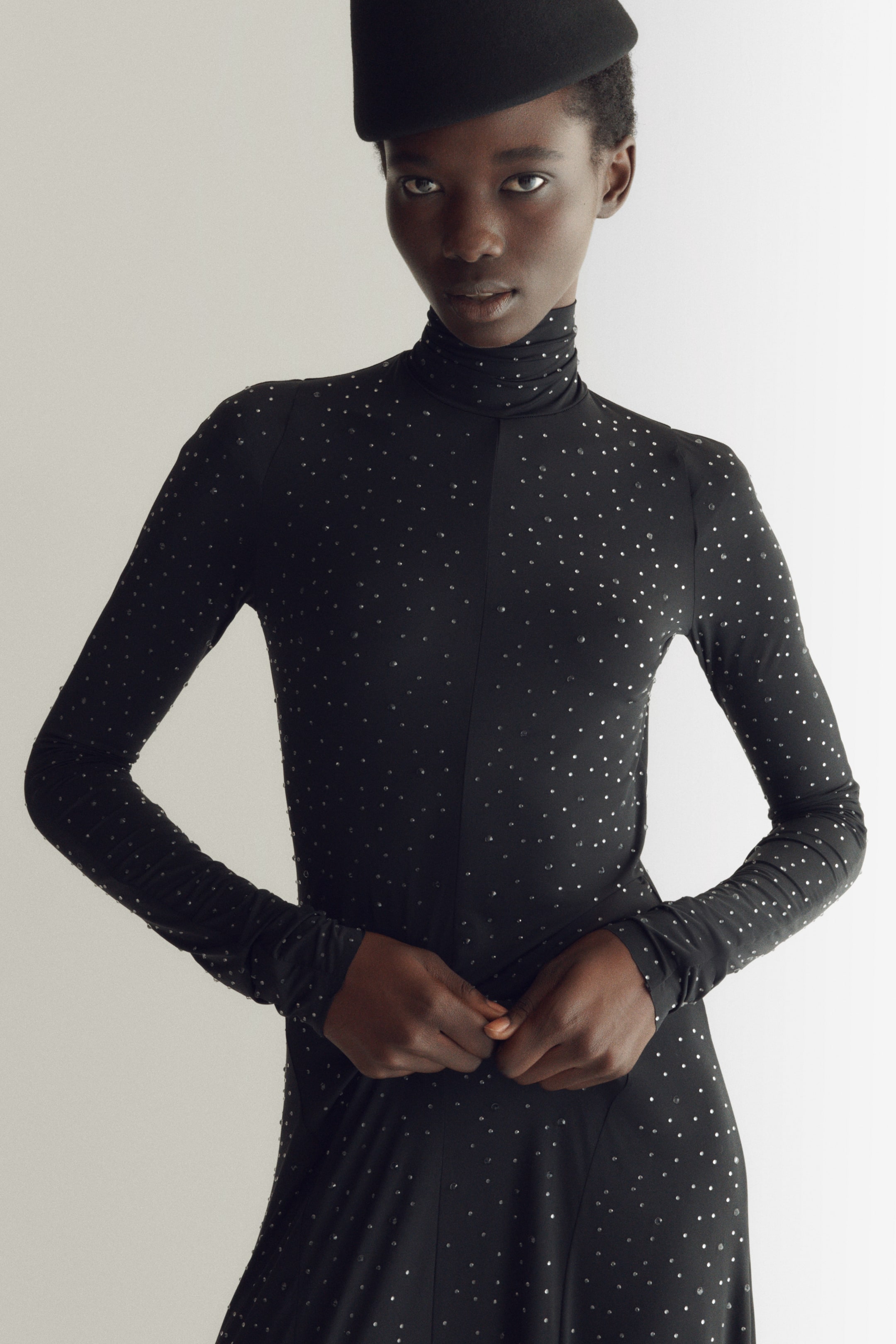 View larger image: A black high-neck dress covered in small, sparkling rhinestones is worn by the model, who holds their hands together at the waist. The dress features long sleeves with a stacked effect at the wrists and a visible center seam, styled with a black structured hat.