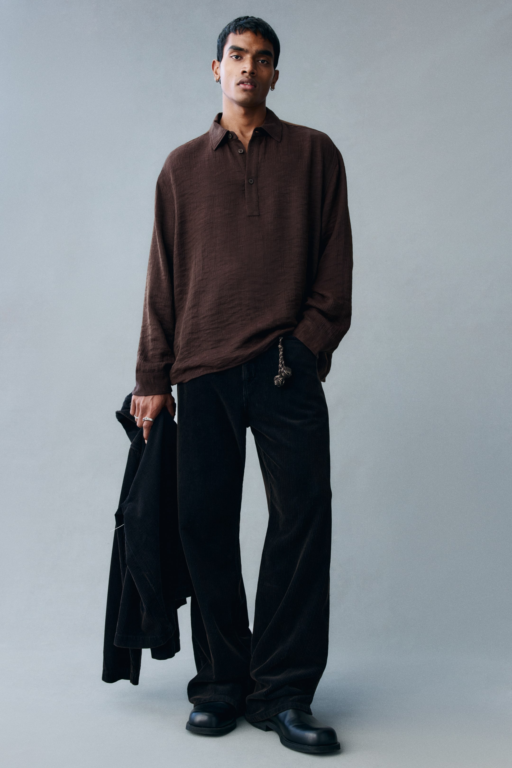 View larger image: The model is wearing a dark brown popover shirt in a crinkled weave, featuring a relaxed fit, a classic collar, and a button placket. The shirt is styled with dark corduroy trousers, and the model is holding a black jacket in one hand.