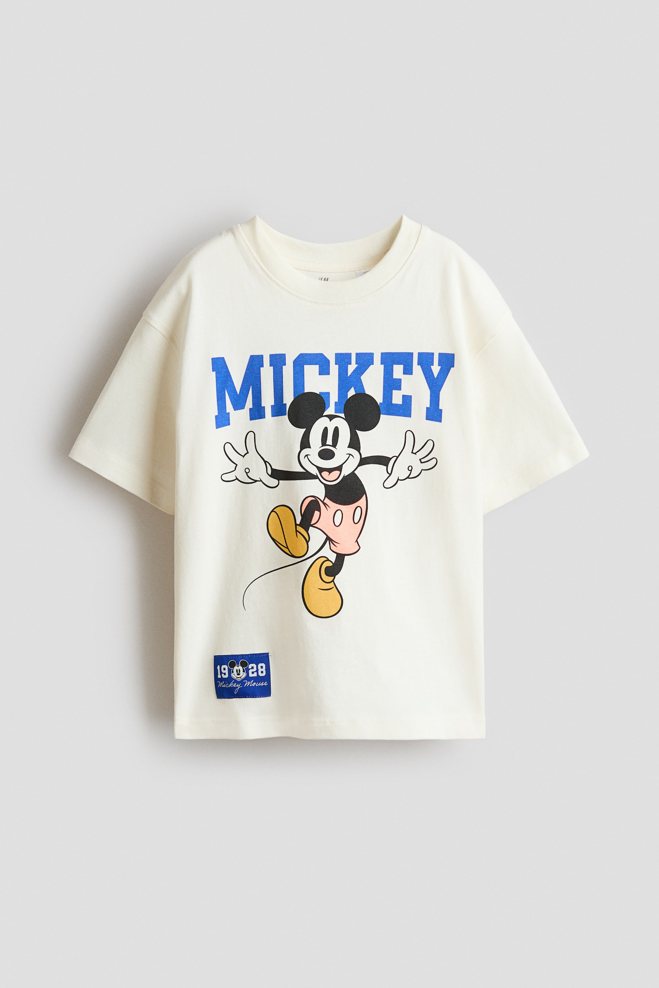View larger image: An oversized T-shirt in soft, printed cotton jersey is presented from the front, in a cream colour with ribbed trim around the neckline and dropped shoulders. The front is adorned with a large graphic print of Mickey Mouse and blue "MICKEY" text, along with a small blue patch reading "19 28 Mickey Mouse" near the bottom-left hem.