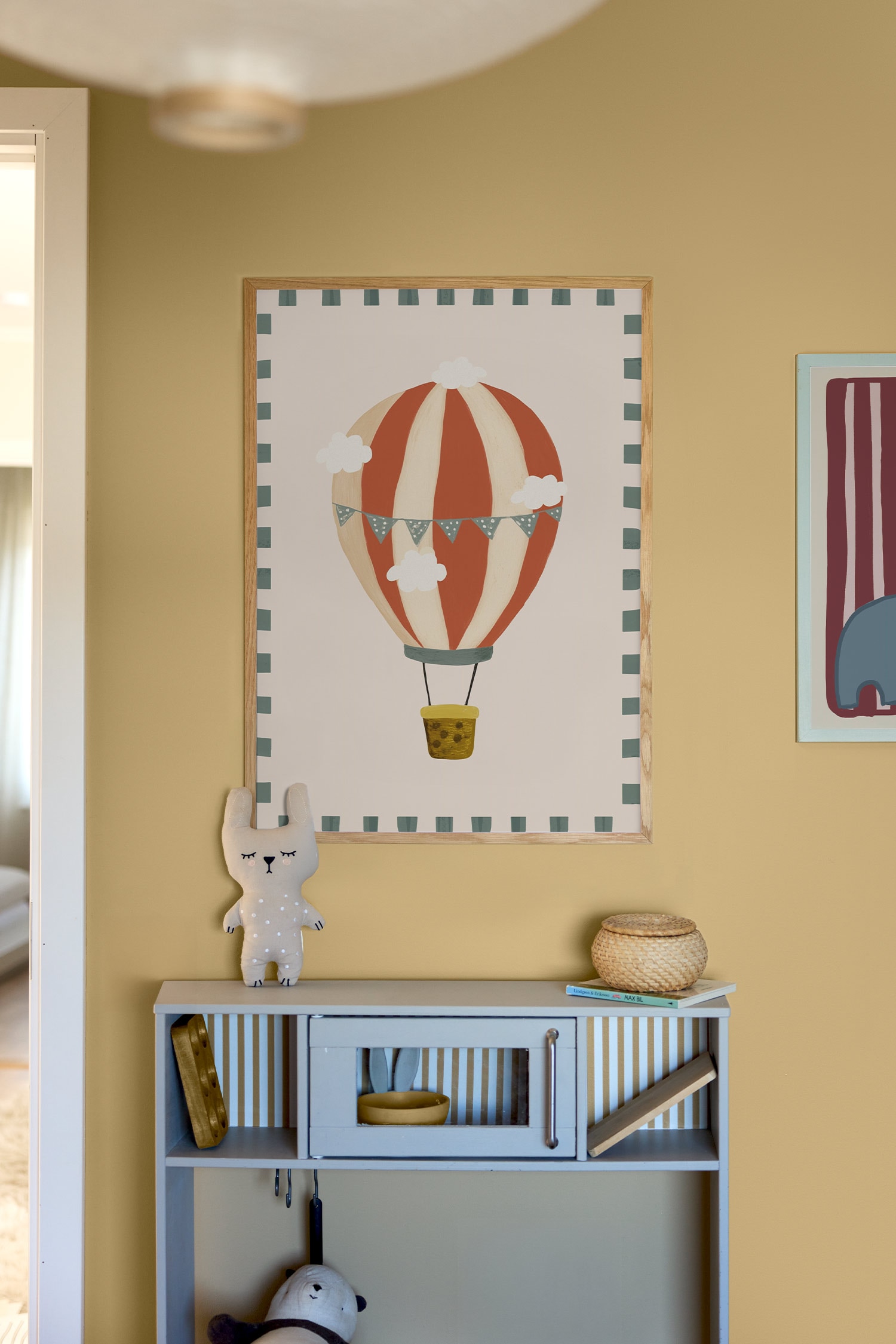 Uma impressão artÃstica apresenta uma ilustração de um balão de ar quente com riscas verticais em vermelho ferrugem e creme, adornado com uma grinalda de galhardetes pontilhados em azul-cinzento e pequenas nuvens brancas. O balão suspende um pequeno cesto amarelo e está disposto sobre um fundo rosa-claro, emoldurado por blocos tracejados verde-escuro.