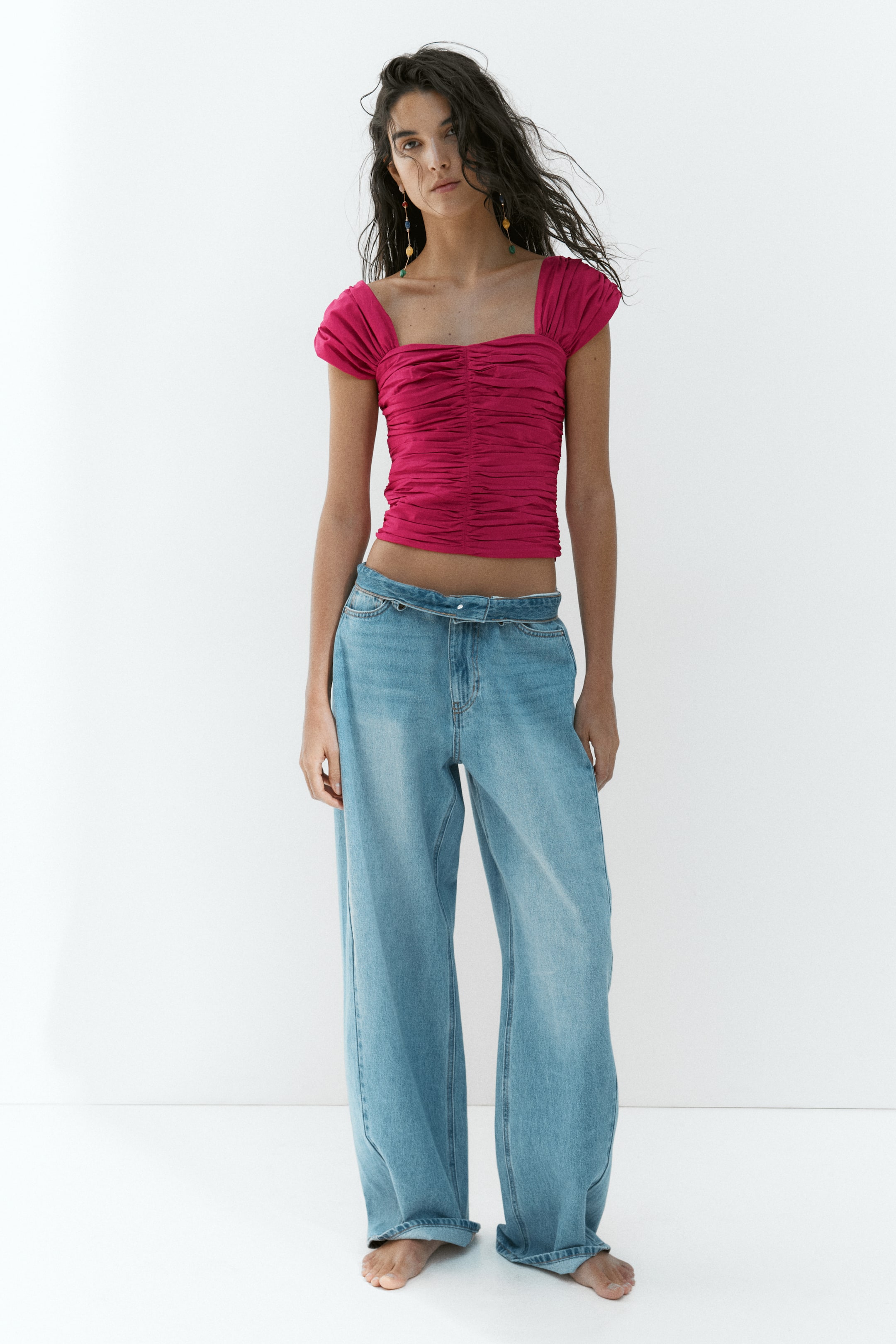Visualizza l'immagine ingrandita: Un top corto aderente color magenta, caratterizzato da uno scollo a cuore, spalline larghe e un ampio arricciamento, è indossato dalla modella, abbinato a jeans in denim a gamba larga con lavaggio chiaro.