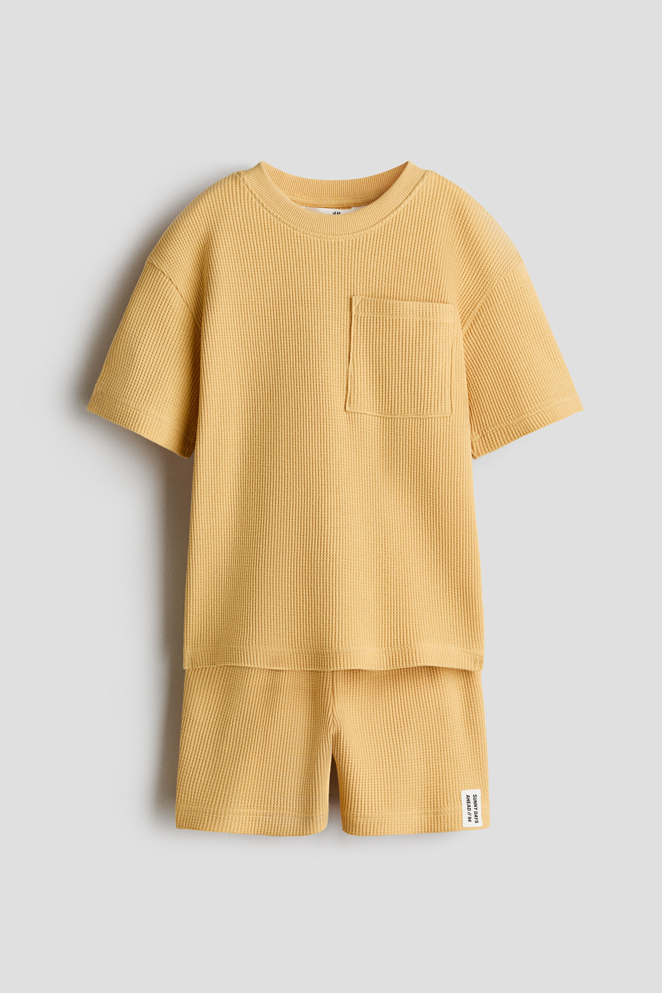Agrandir l'image: Un ensemble en jersey de coton gaufré jaune comprenant un T-shirt et un short assorti. Le T-shirt présente un col rond avec bordure côtelée, une poche poitrine plaquée à gauche et des épaules tombantes, tandis que le short affiche une taille élastiquée avec cordon de serrage et une petite étiquette blanche sur la jambe droite.