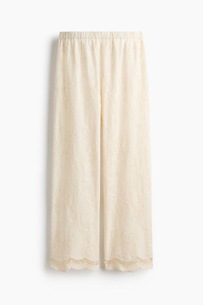 Pantalonii de culoare crem sunt prezentați cu fața în față, având o talie elasticizată. Aceștia prezintă un model de dantelă florală pe toată suprafața și se termină cu un tiv din dantelă ondulată la margini. - 1
