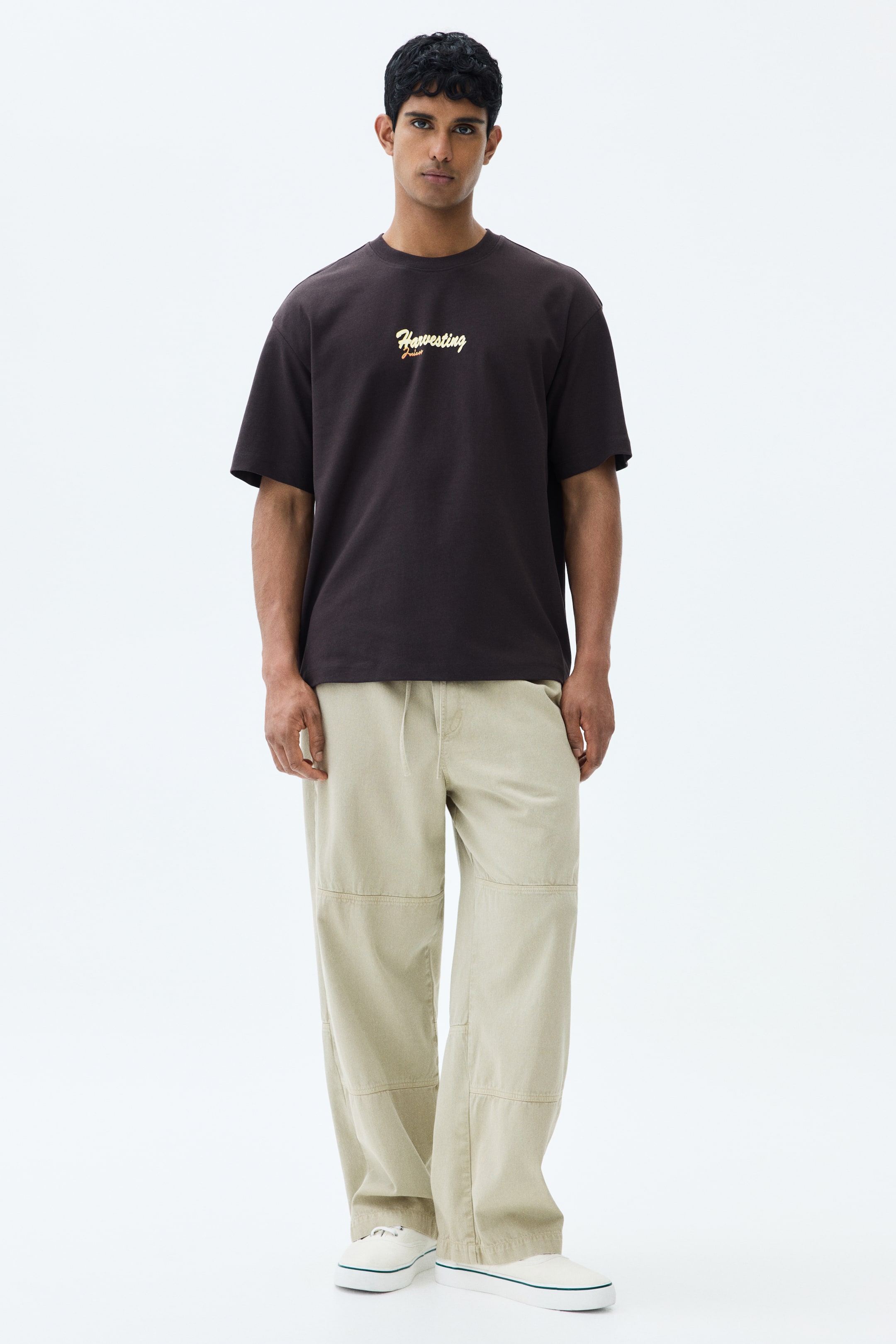 View larger image: The model is donning light beige trousers crafted from a cotton weave. These trousers boast a covered elasticated waist complemented by a visible drawstring, diagonal side pockets, and unique paneling on the legs, offering a relaxed fit.
