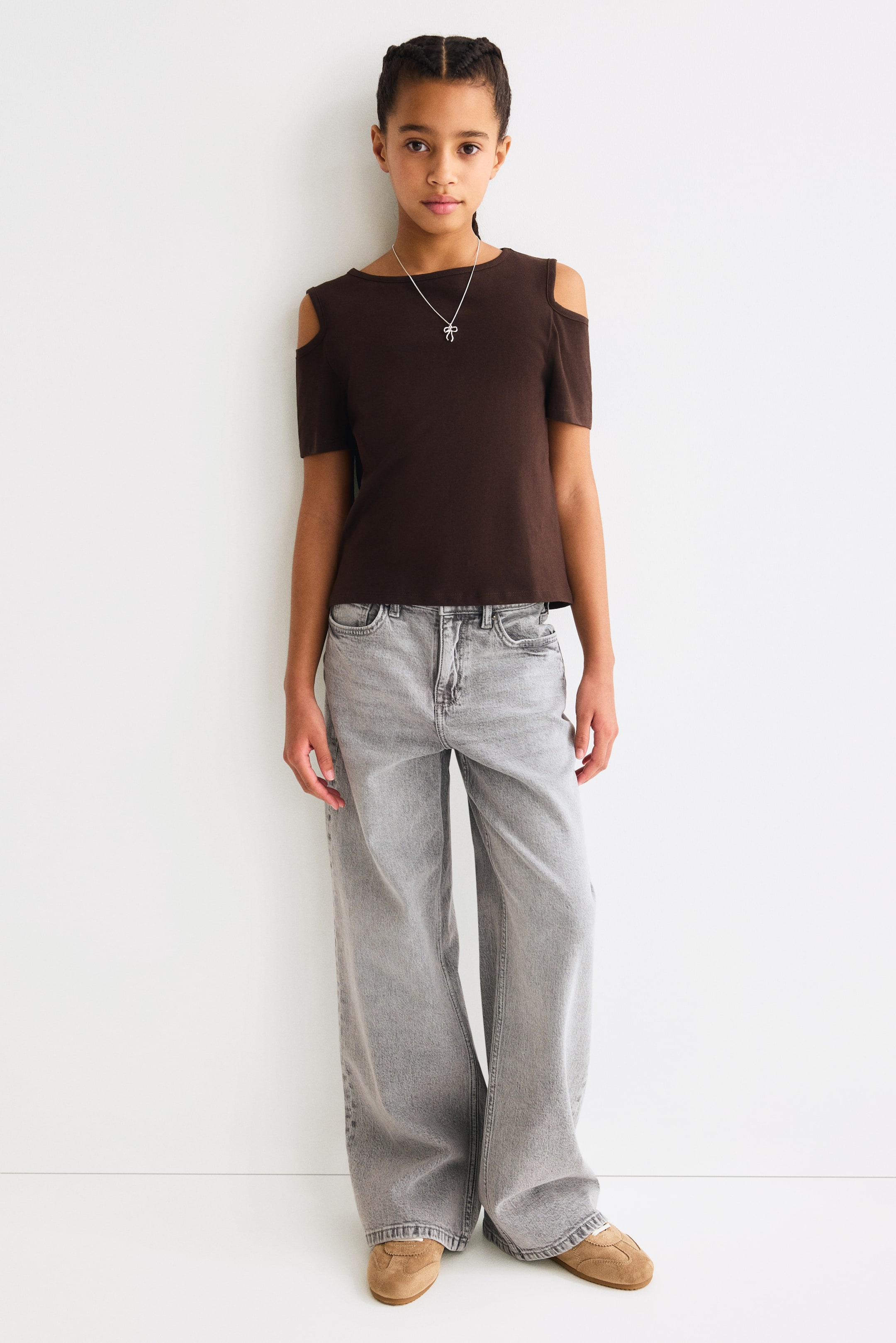 Ver imagen más grande: La blusa marrón oscuro con hombros descubiertos presenta un escote redondo y mangas cortas, con aberturas visibles en los hombros. Un collar plateado con un colgante de lazo descansa sobre la blusa.