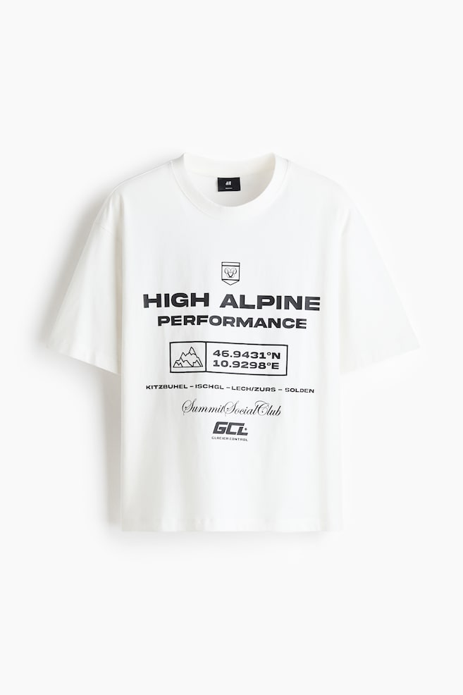 A white crew neck t-shirt is shown from the front, featuring a relaxed fit and wide short sleeves. The chest is displaying a black graphic print that includes the bold text "HIGH ALPINE PERFORMANCE" along with geographic coordinates, a mountain graphic, and alpine-themed text. - 1