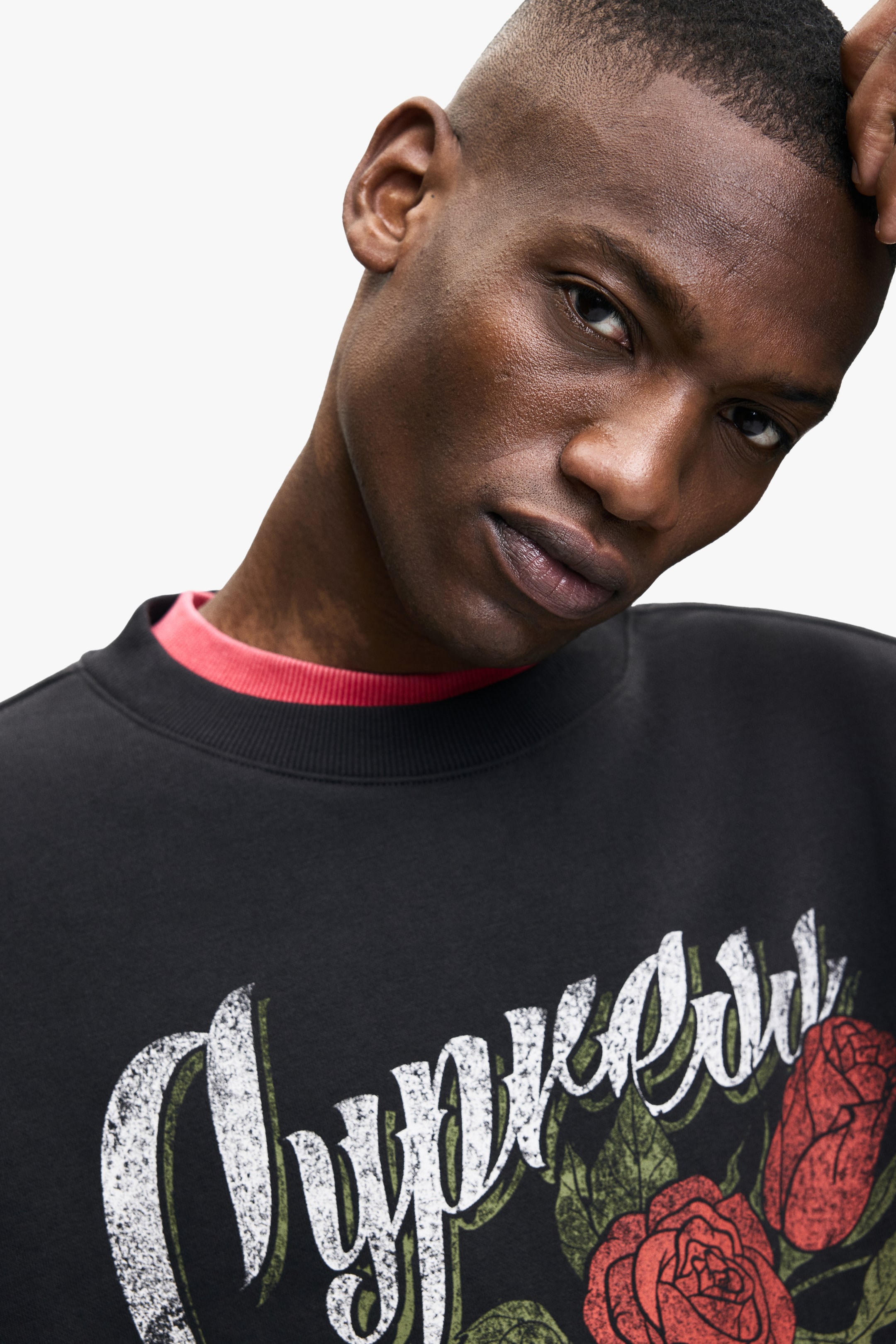 View larger image: A black crew-neck sweatshirt, featuring a distressed white script graphic with red roses and green leaves on the front, is being worn by the model. A red ribbed mock neck t-shirt is being layered underneath, visible at the collar.