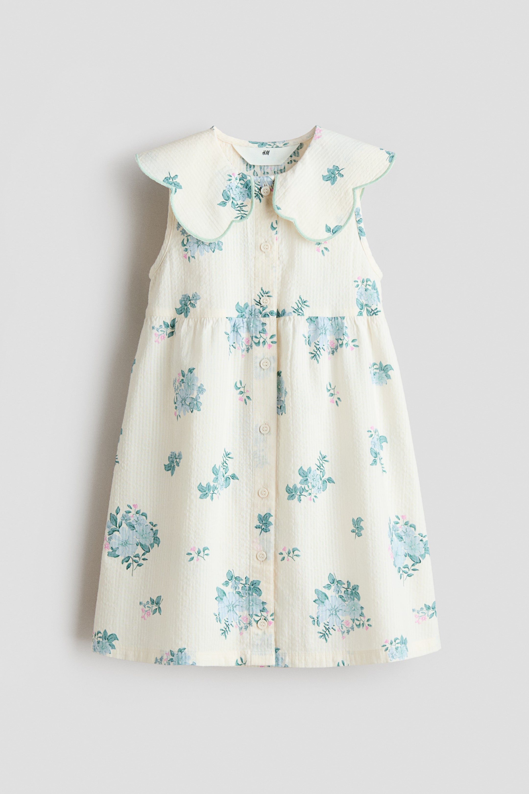 Büyük resmi görüntüle: Kolsuz bir elbise ÖNDEN sunulmaktadır, krem rengi, hafif dokulu kumaşı üzerine dağılmış mavi ve yeşil çiçek desenleri içermektedir. Hafif yeşil biyeyle çevrelenmiş büyük, dalgalı bir Peter Pan yakaya, krem renkli düğmelerle tam boy düğme plaketine sahiptir ve rahat, A-kesim bir silüet oluşturmak için bele doğru büzülmektedir.