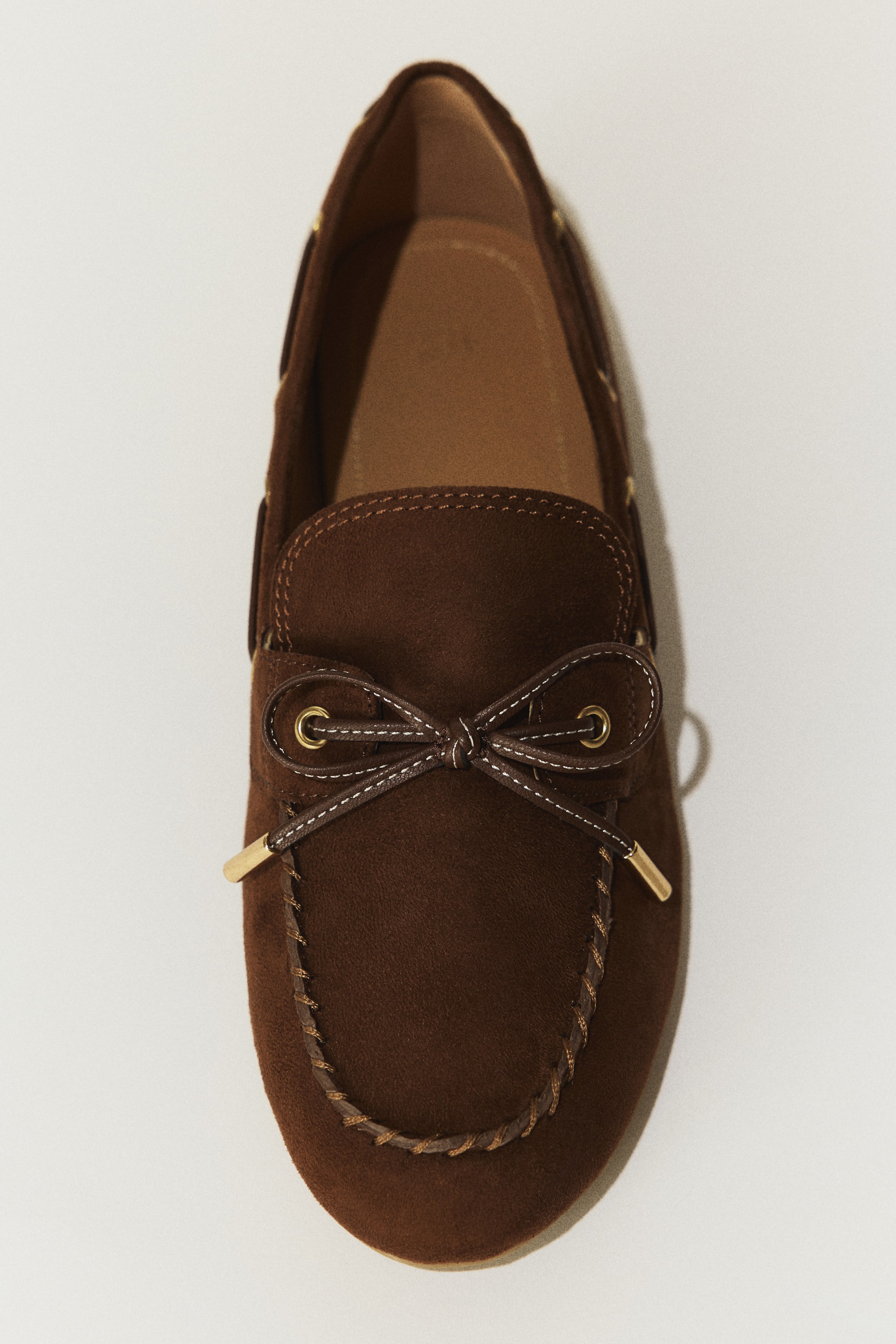 Größeres Bild ansehen: Ein dunkelbrauner Wildleder-Mokassin-Slipper ist aus der Draufsicht zu sehen; er besitzt eine dekorative Schleife, gebunden mit kontrastierenden hellbraunen Schnürsenkeln mit goldfarbenen Aglets und Eyelets, und eine geflochtene Borte umrundet das Moc-Toe.