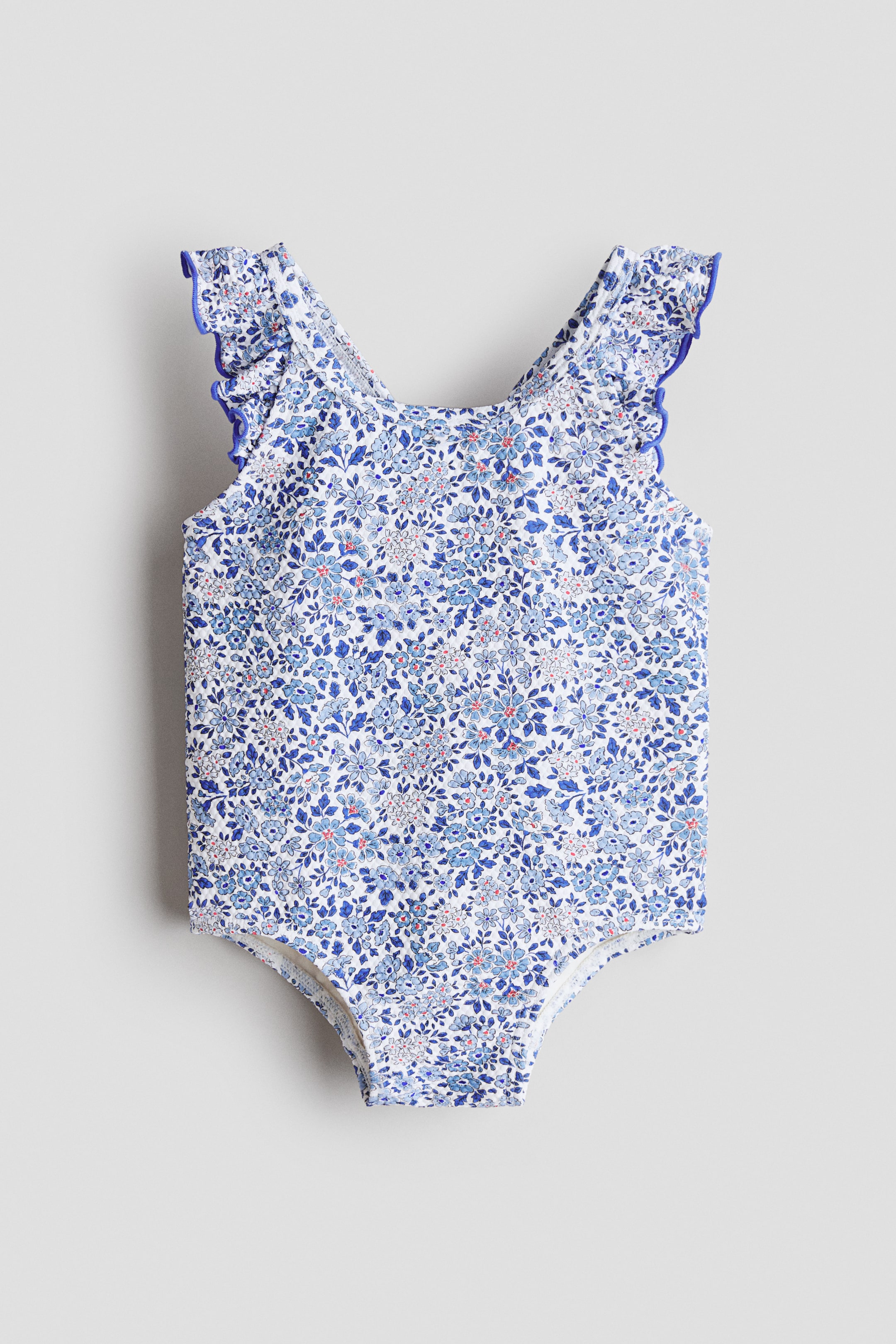 Zobacz większy obraz: Biały jednoczęściowy kostium kąpielowy prezentuje PRZÓD, z gęstym kwiatowym wzorem w różnych odcieniach niebieskiego z drobnymi czerwonymi akcentami. Posiada falbankowane ramiączka, każde obszyte kontrastową niebieską lamówką.