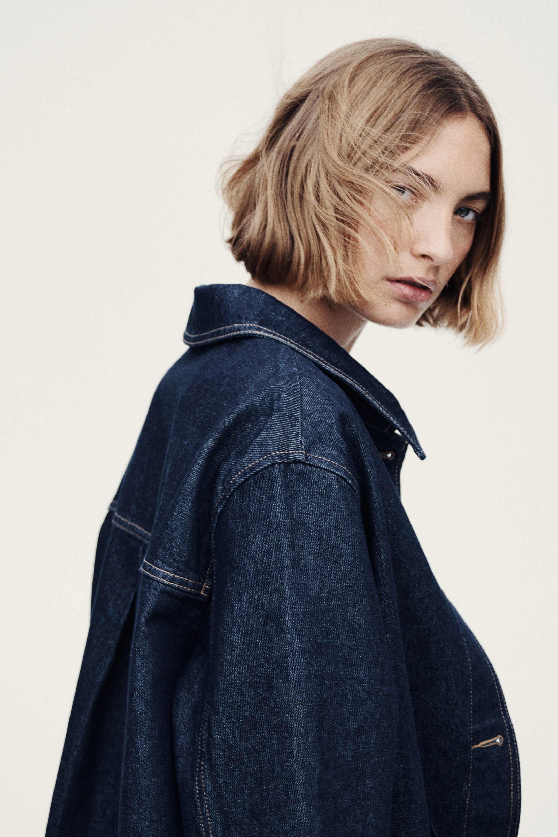View larger image: A dark blue denim jacket is displayed from a side angle, showcasing a structured pointed collar and tan contrast stitching. The back yoke, a vertical pleat on the back, and snap buttons on the partially open front placket are visible.