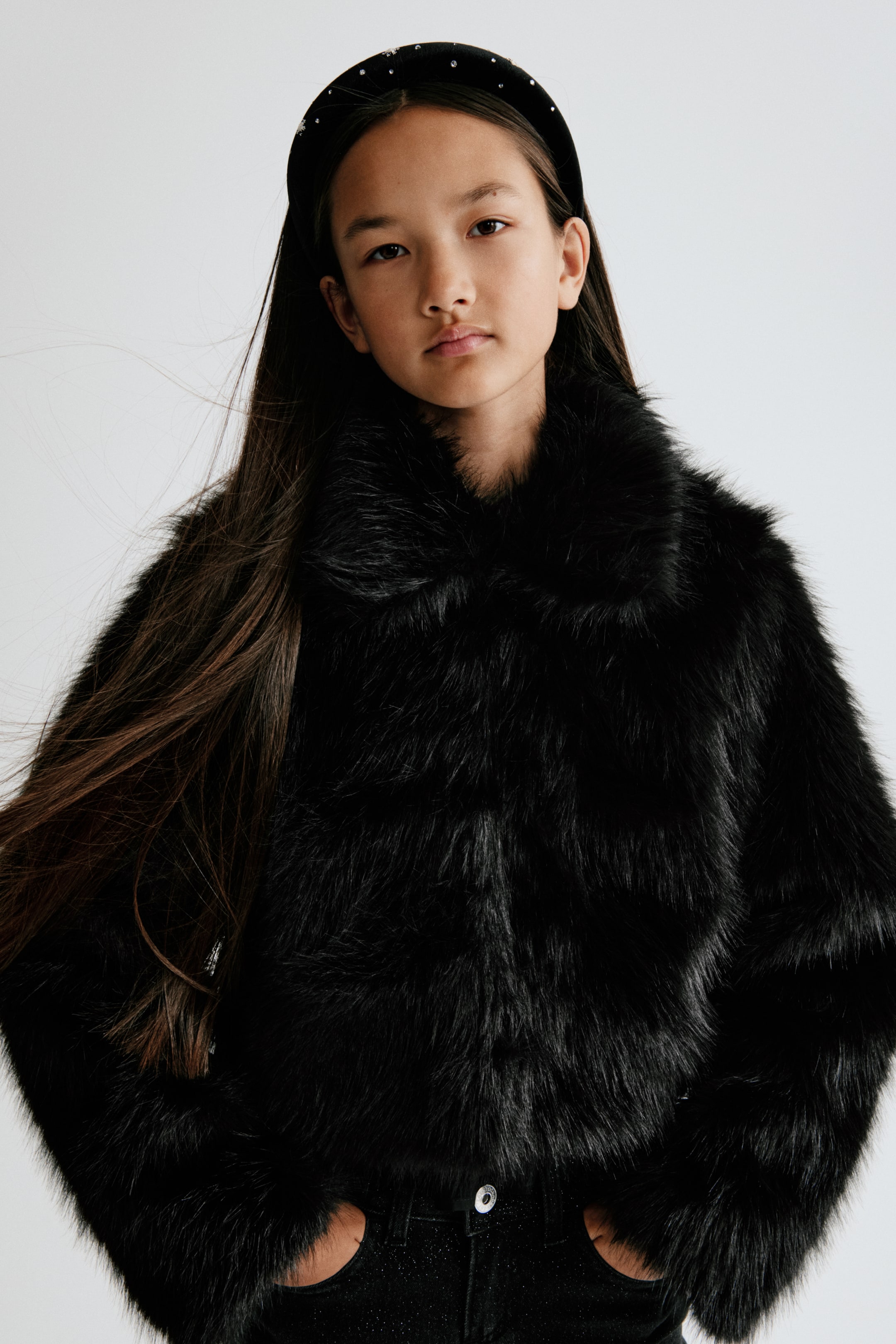 View larger image: A short, fluffy black faux-fur jacket is shown from the front, featuring a wide collar and a closed-front design. The jacket is styled with black jeans, highlighting its waist-length cut and relaxed fit.
