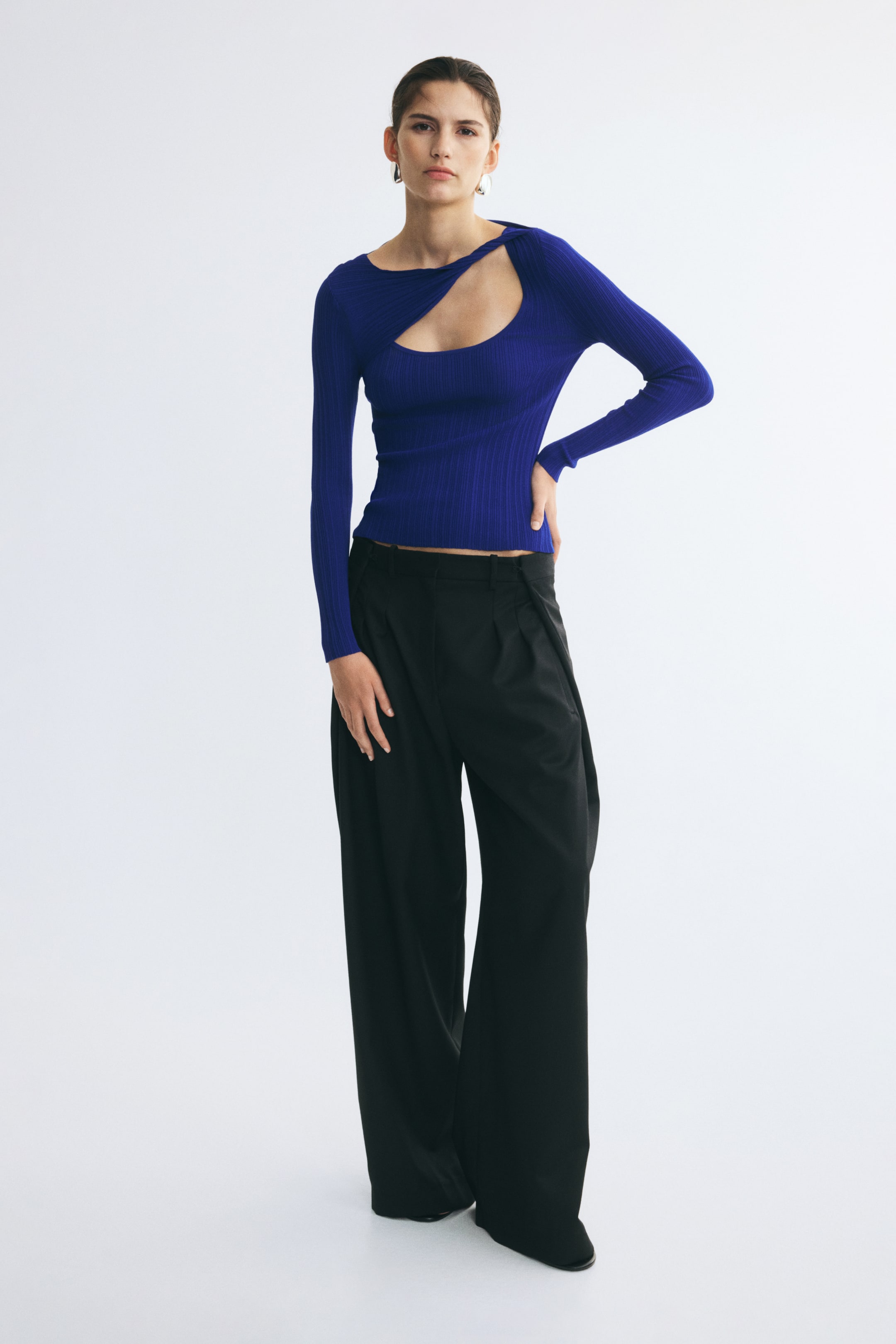 Grotere afbeelding bekijken: Een levendigblauwe top in ribgebreide stof met een asymmetrische halslijn, een gedraaid cut-outdetail op de borst en lange mouwen wordt gedragen door het model. De aansluitende top is gestyled met een zwarte broek met wijde pijpen.