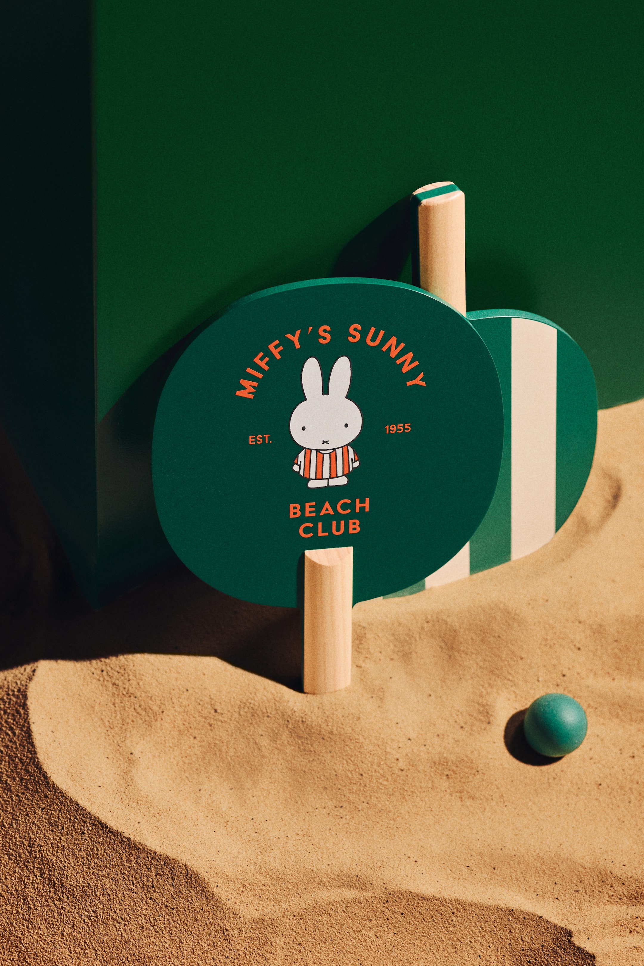 Agrandir l'image: Deux raquettes de plage avec des manches en bois clair sont plantées verticalement dans le sable, accompagnées d'une petite balle turquoise. La raquette avant est vert foncé, arborant un personnage blanc de Miffy en chemise rayée et le texte orange 'Miffy's Sunny Beach Club Est. 1955', tandis que la deuxième raquette partiellement visible est vert foncé avec une bande blanche verticale.