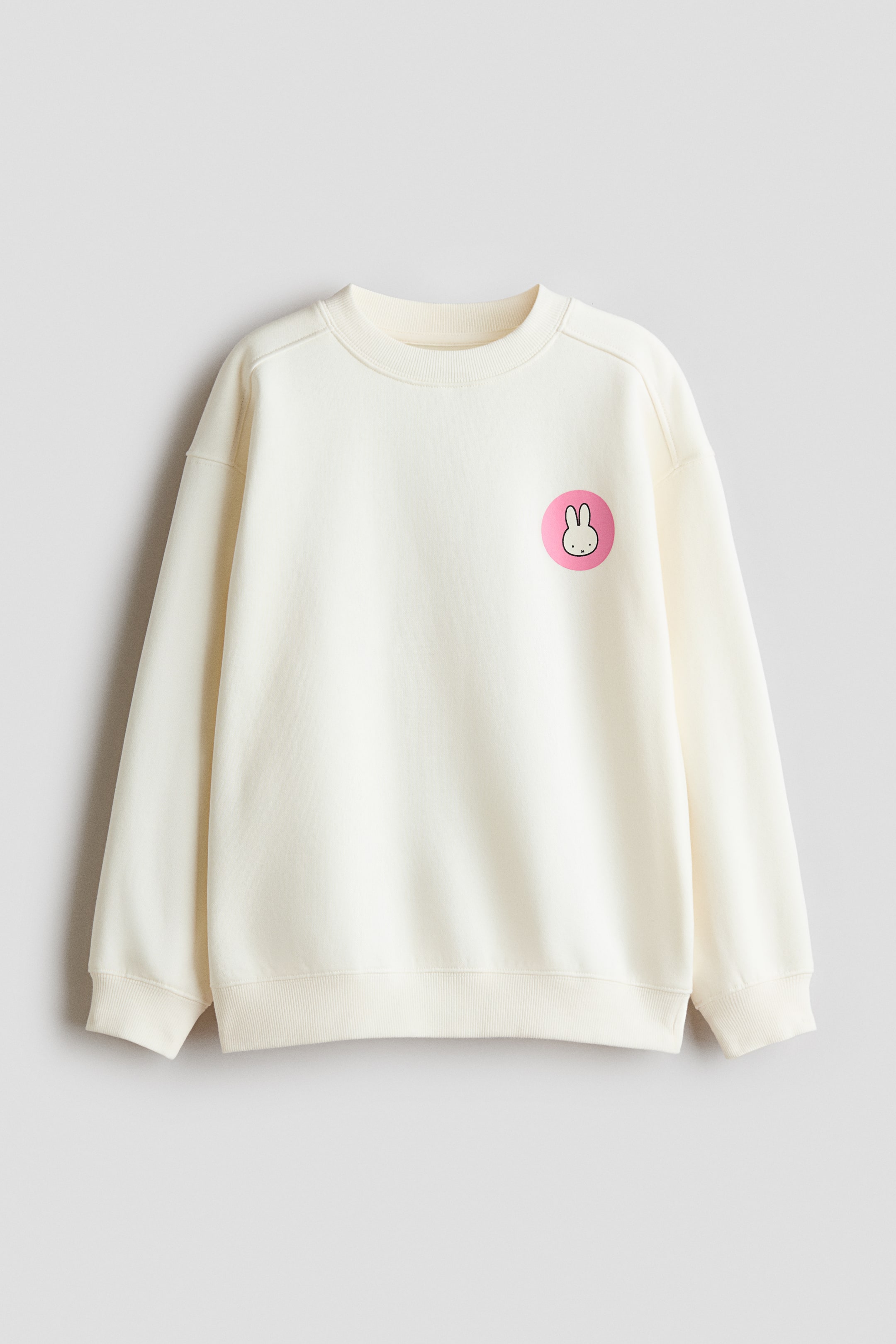 Größeres Bild ansehen: Ein cremefarbenes Sweatshirt in lockerer Passform ist von vorn zu sehen und zeigt auf der linken Brust ein kleines, rundes pinkfarbenes Grafikmotiv eines Zeichentrickkaninchens. Das Modell verfügt über einen gerippt eingefassten Rundhalsausschnitt, überschnittene Schultern sowie gerippte Bündchen und einen gerippten Saum.