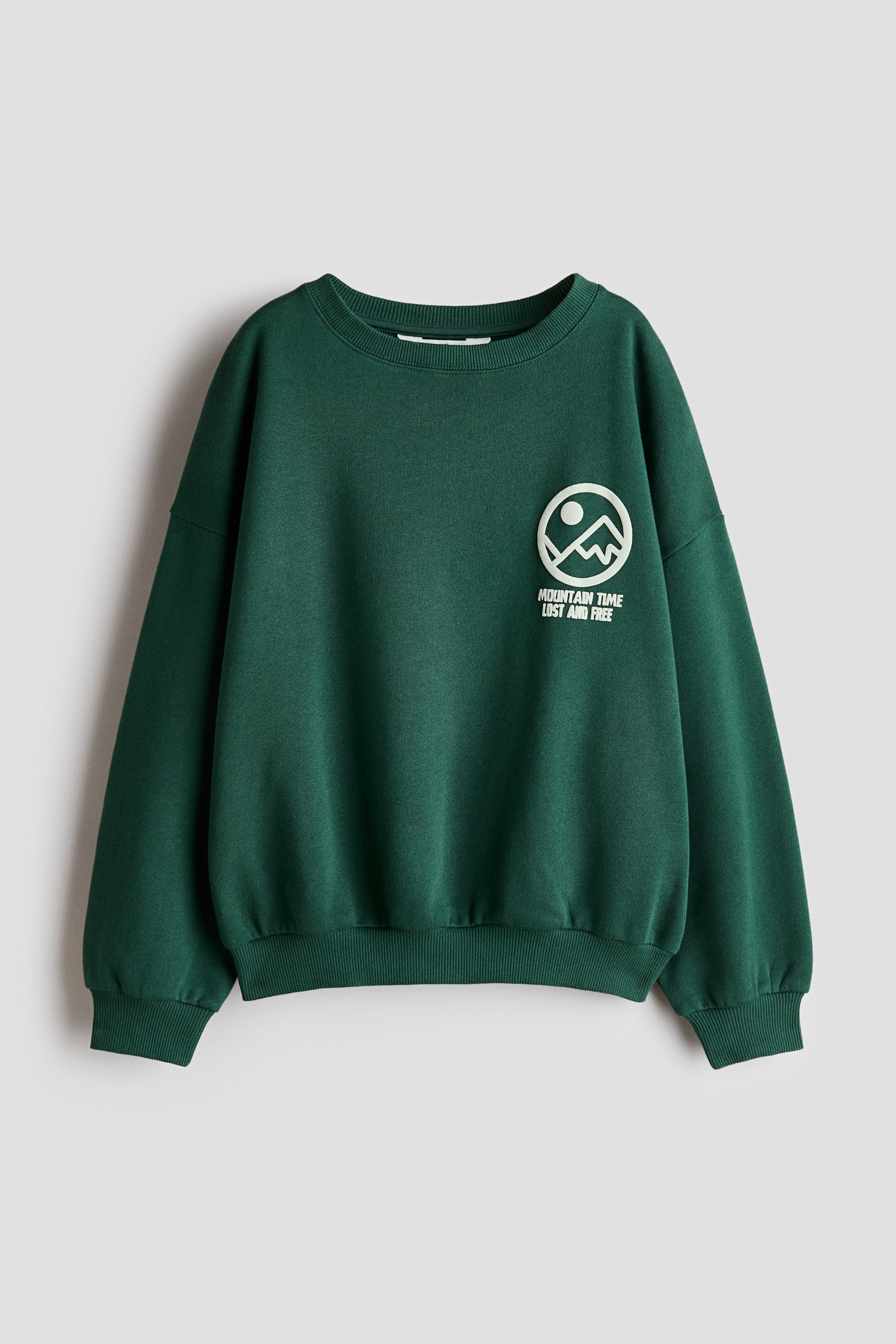 View larger image: A dark green crew neck sweatshirt is shown from the front, featuring dropped shoulders, ribbed cuffs, and a ribbed hem. A circular white graphic depicting a mountain range and sun, with the text "MOUNTAIN TIME LOST AND FREE" printed beneath, is positioned on the left chest.