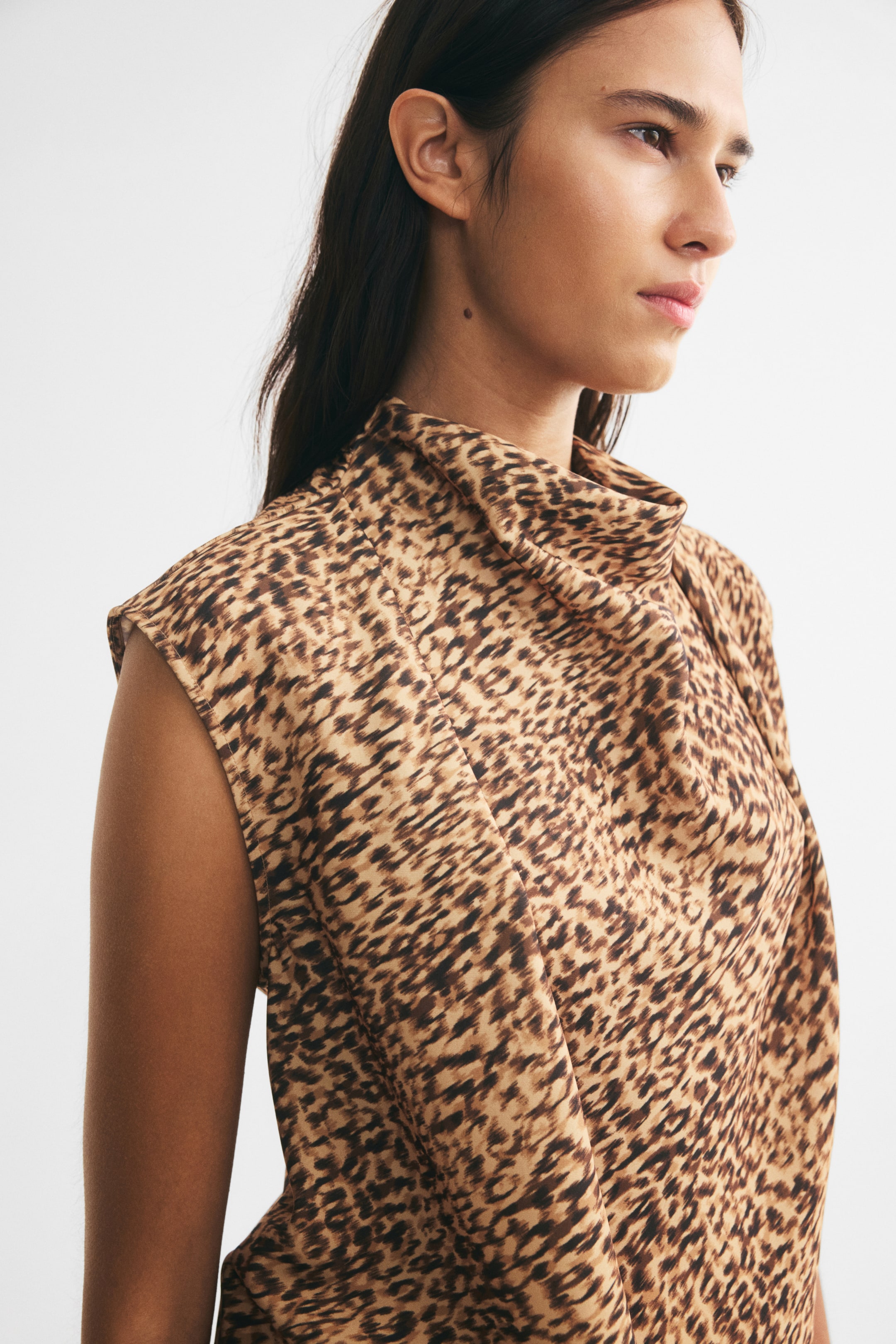 View larger image: The model is wearing a sleeveless leopard-print top with a draped cowl neckline and shoulder pleats. Shown from the side, the top highlights its woven fabric with a brown and black animal pattern.