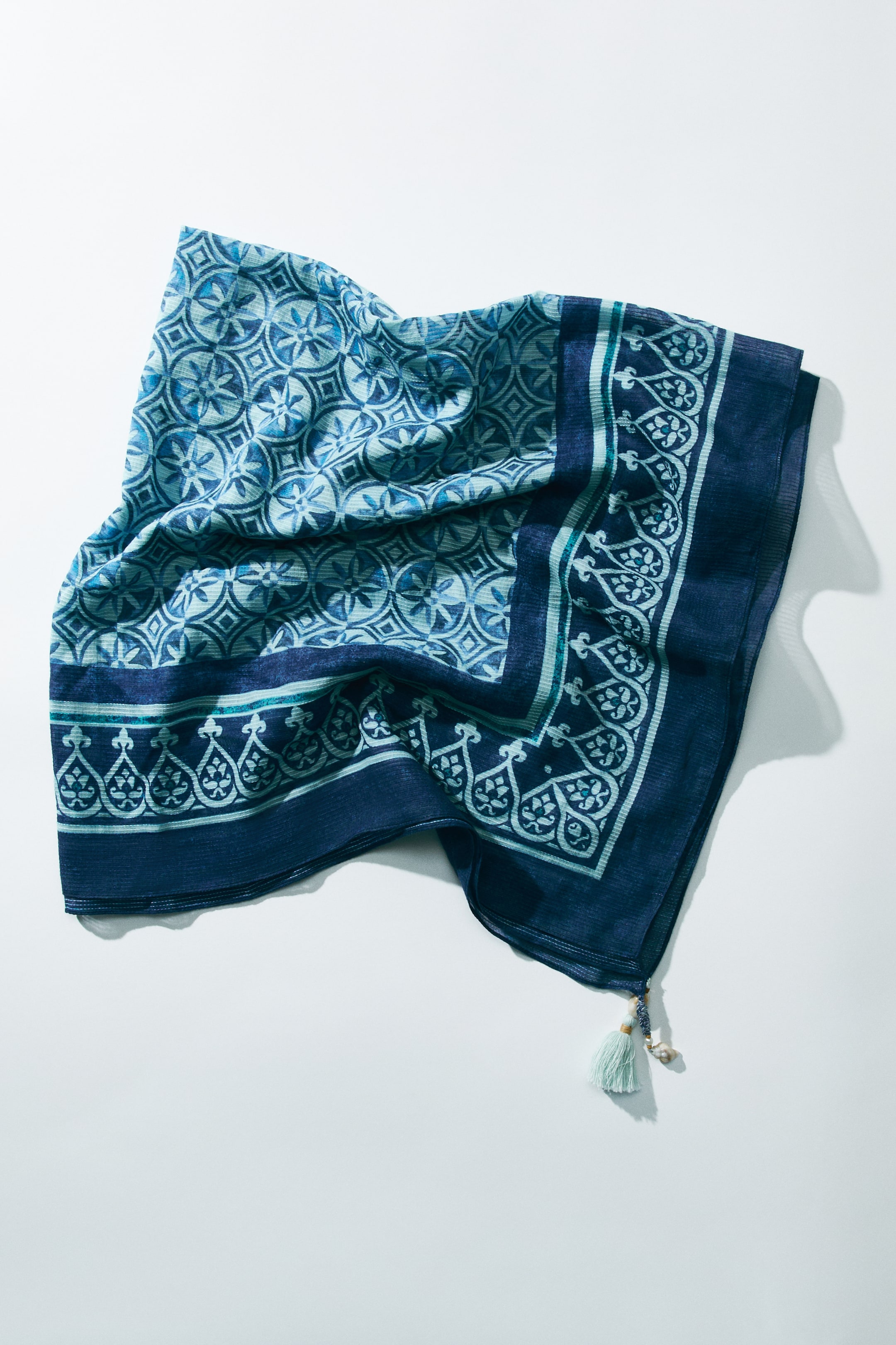 Ver imagen más grande: Un pañuelo cuadrado de tejido de algodón presenta un patrón central de motivos circulares y un borde con diseños intrincados en varios tonos de azul. Un borlón azul claro y un pequeño adorno en forma de concha marina detallan una esquina.