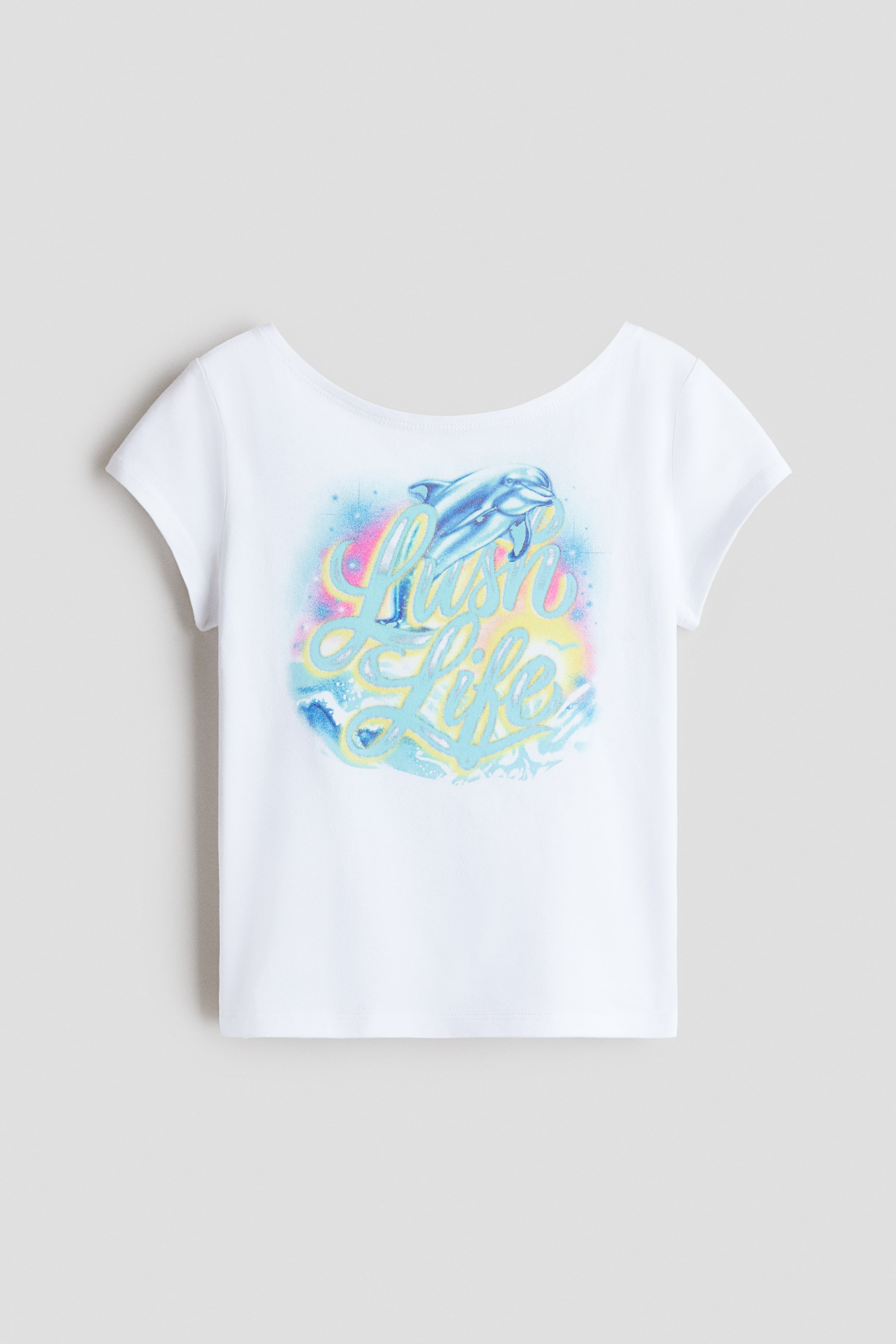 View larger image: The white t-shirt showcases a wide, rounded neckline and short cap sleeves. A vibrant graphic is centered on the front, illustrating a blue dolphin soaring above the script text "Lush Life," encircled by pink, yellow, and blue spray-paint effects and wave patterns.