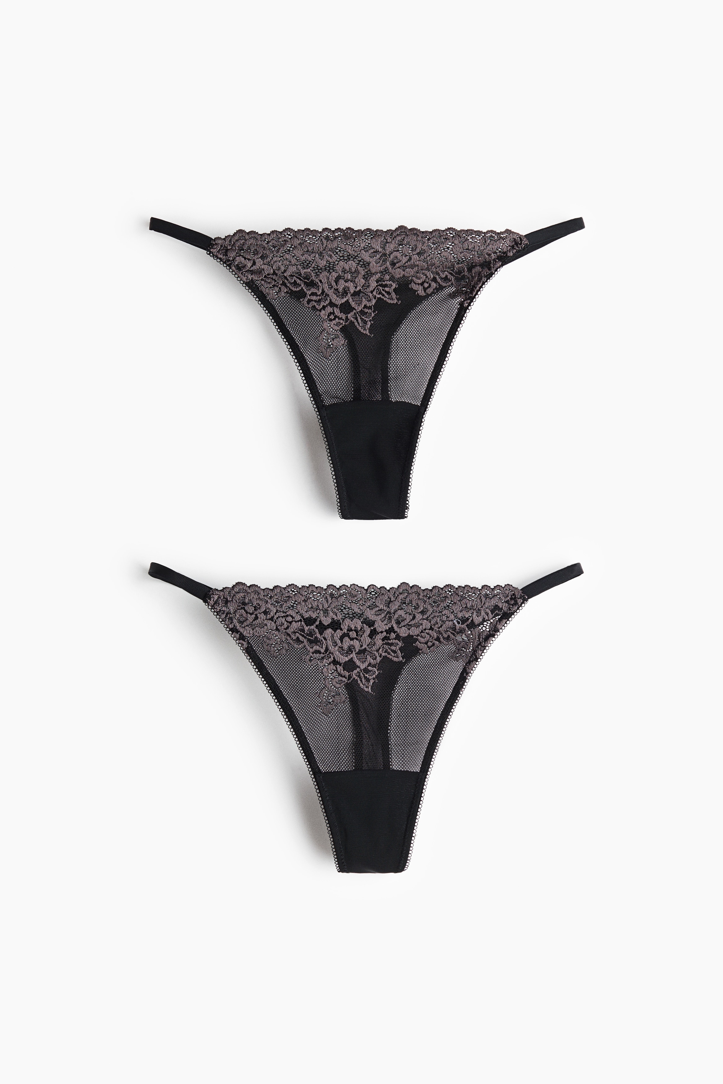 The model is wearing a black mesh G-string with a low-rise waist, dark grey lace trim and narrow sides. The G-string is paired with a matching black underwired bra, decorated with white lace overlays.