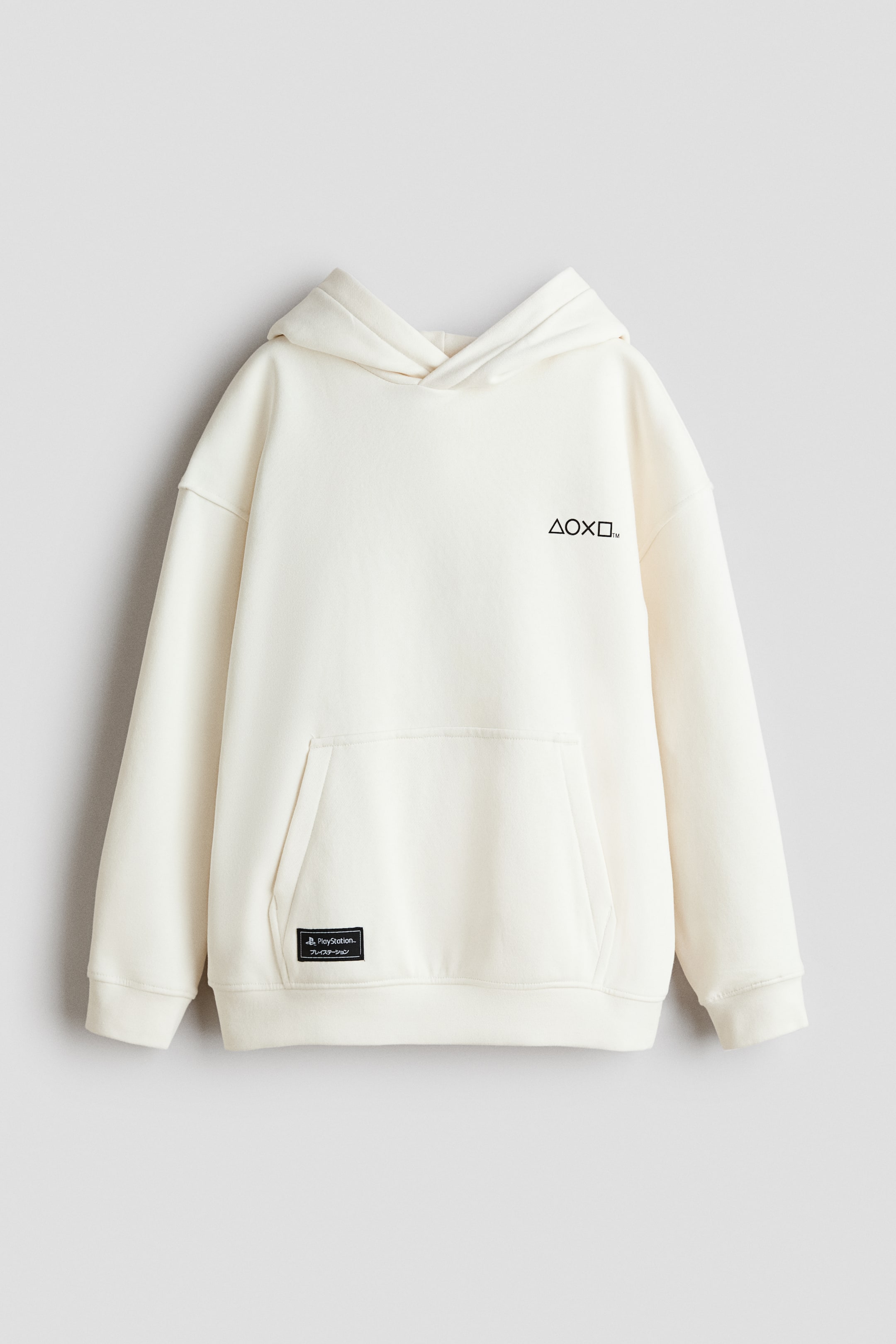 View larger image: A cream-colored hoodie is shown from the front, showcasing a black graphic symbol on the upper chest and a large kangaroo pocket on the lower front. A small black rectangular patch with the text "PlayStation." is visible on the left side of the pocket, and ribbed cuffs and hem are included.