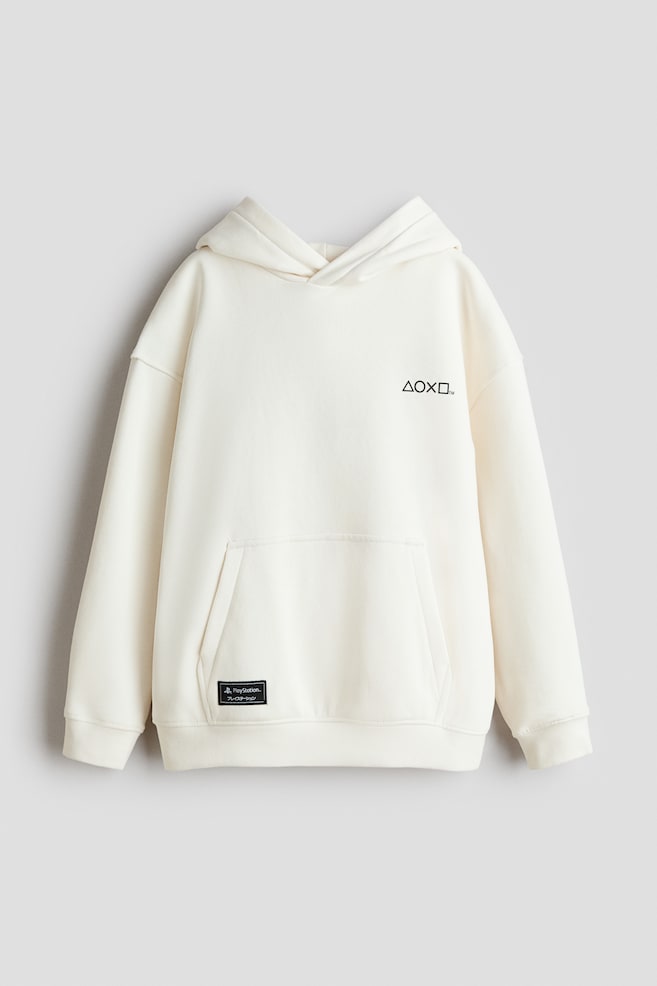 A cream-coloured hoodie is showcased from the front, featuring a black graphic symbol on the upper chest and a large kangaroo pocket on the lower front. A small black rectangular patch with "PlayStation." text is visible on the left side of the pocket, and ribbed cuffs and hem are present. - 1