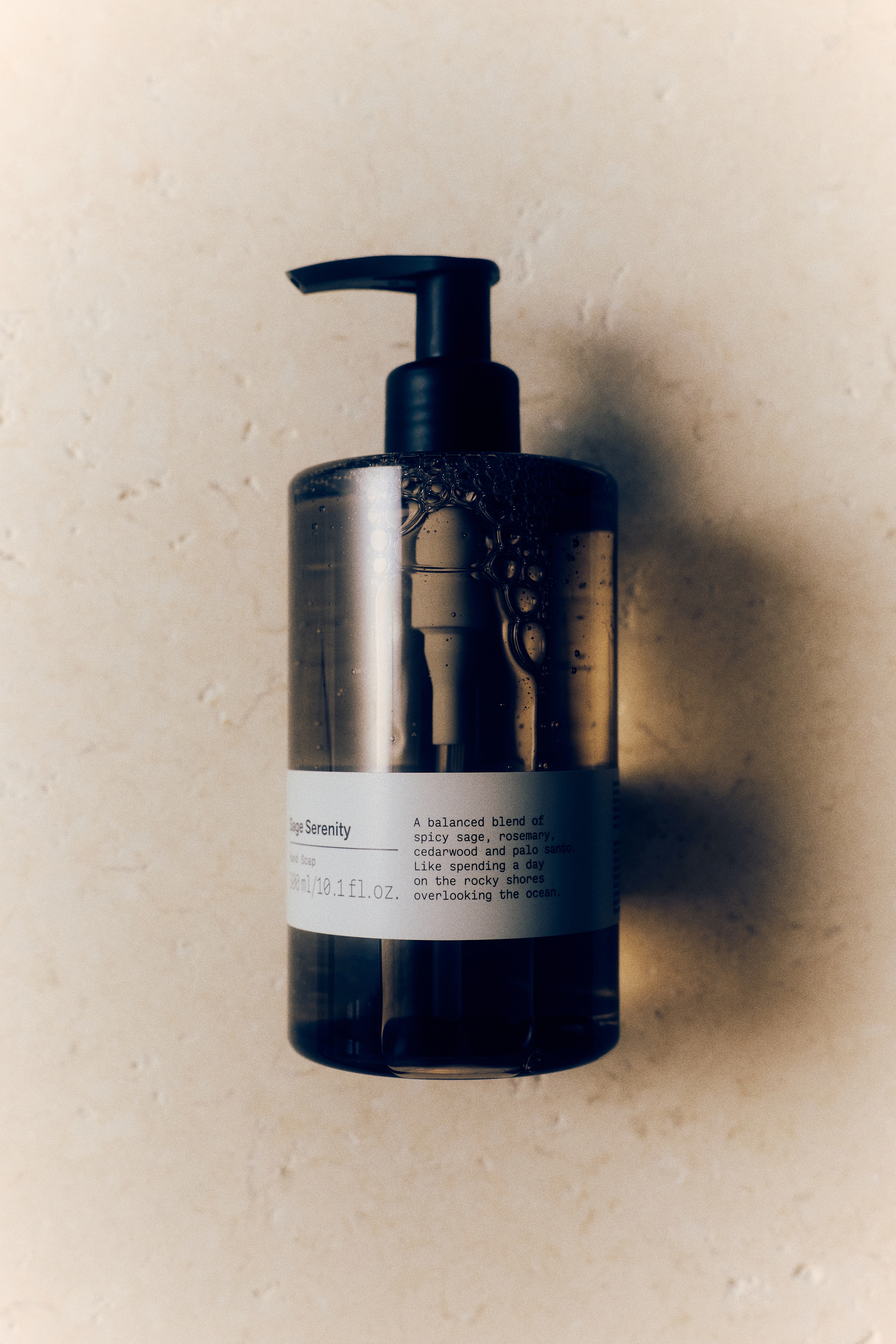 A cylindrical, translucent brown bottle of Sage Serenity hand soap is standing upright, facing front. It is featuring a black pump dispenser on top and is displaying a clean white label with black text on its lower portion.