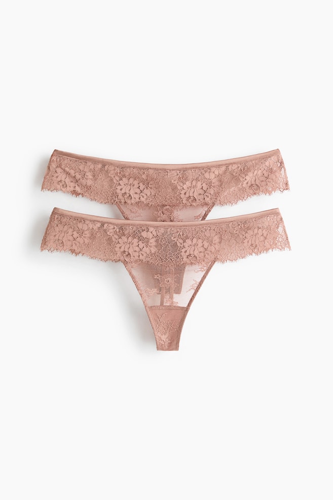 "Duas tangas em rosa empoeirado estão deitadas planas, ambas voltadas para a frente e ligeiramente sobrepostas. São feitas de renda floral com bordas recortadas e malha transparente." - 1