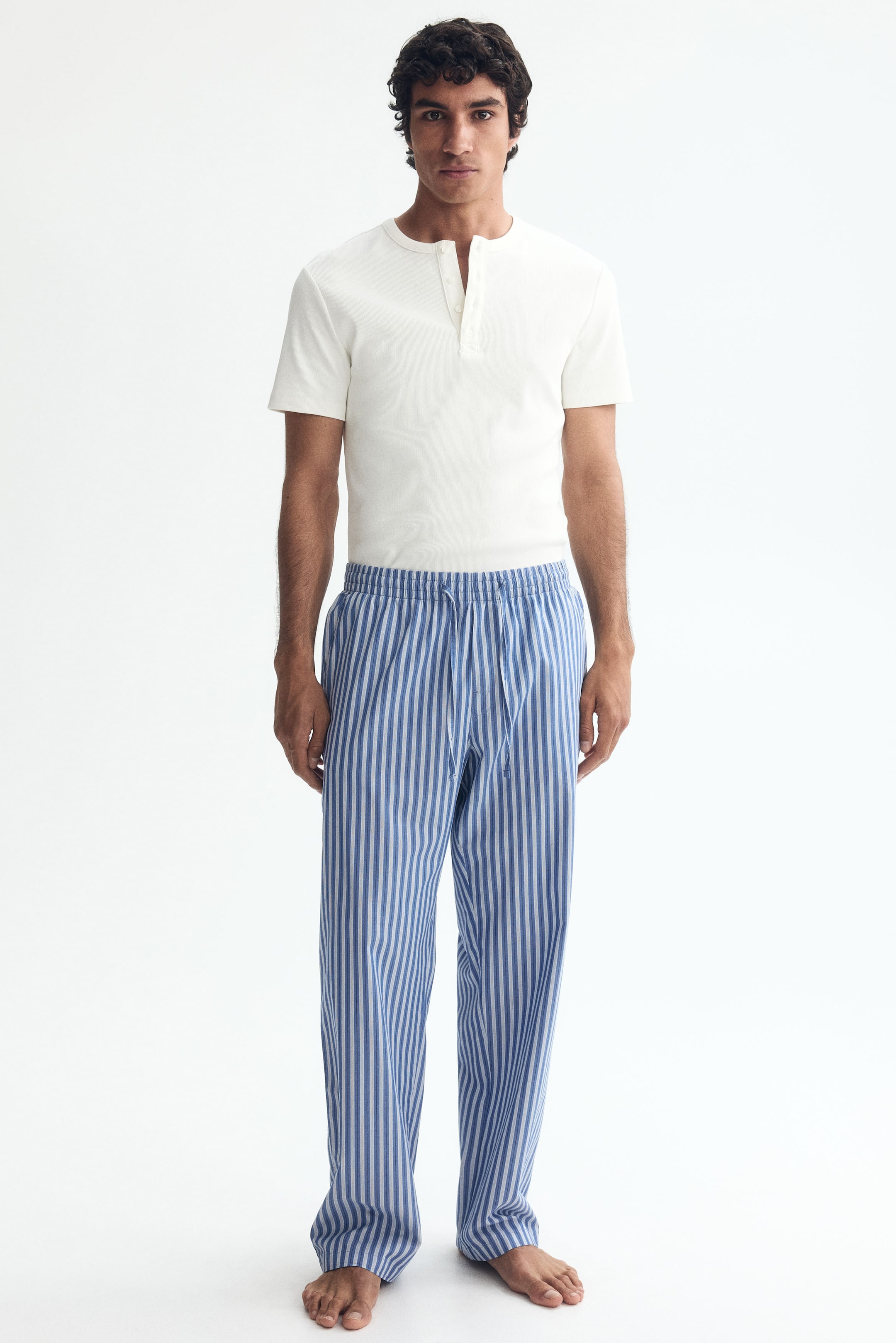 View larger image: The model is showcasing blue and white vertically striped trousers, designed with an elasticated waistband and a tied drawstring. These trousers feature visible side pockets and offer a relaxed fit.