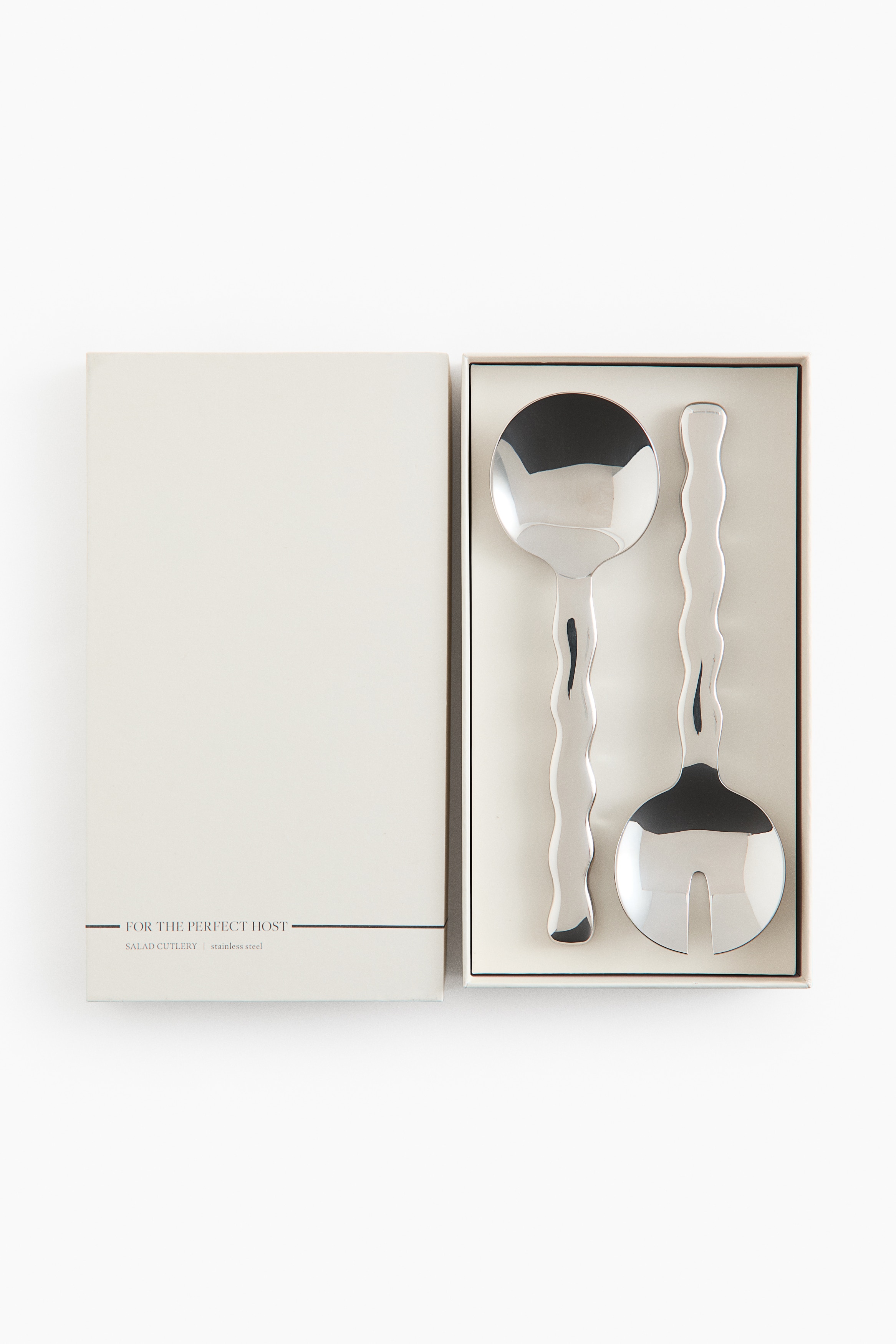 Polished stainless steel salad servers featuring wavy, undulating handles are resting side by side inside a gift box lined with crumpled white tissue paper. The set includes a serving spoon and a serving fork with a central slot, both showcasing a highly reflective finish.
