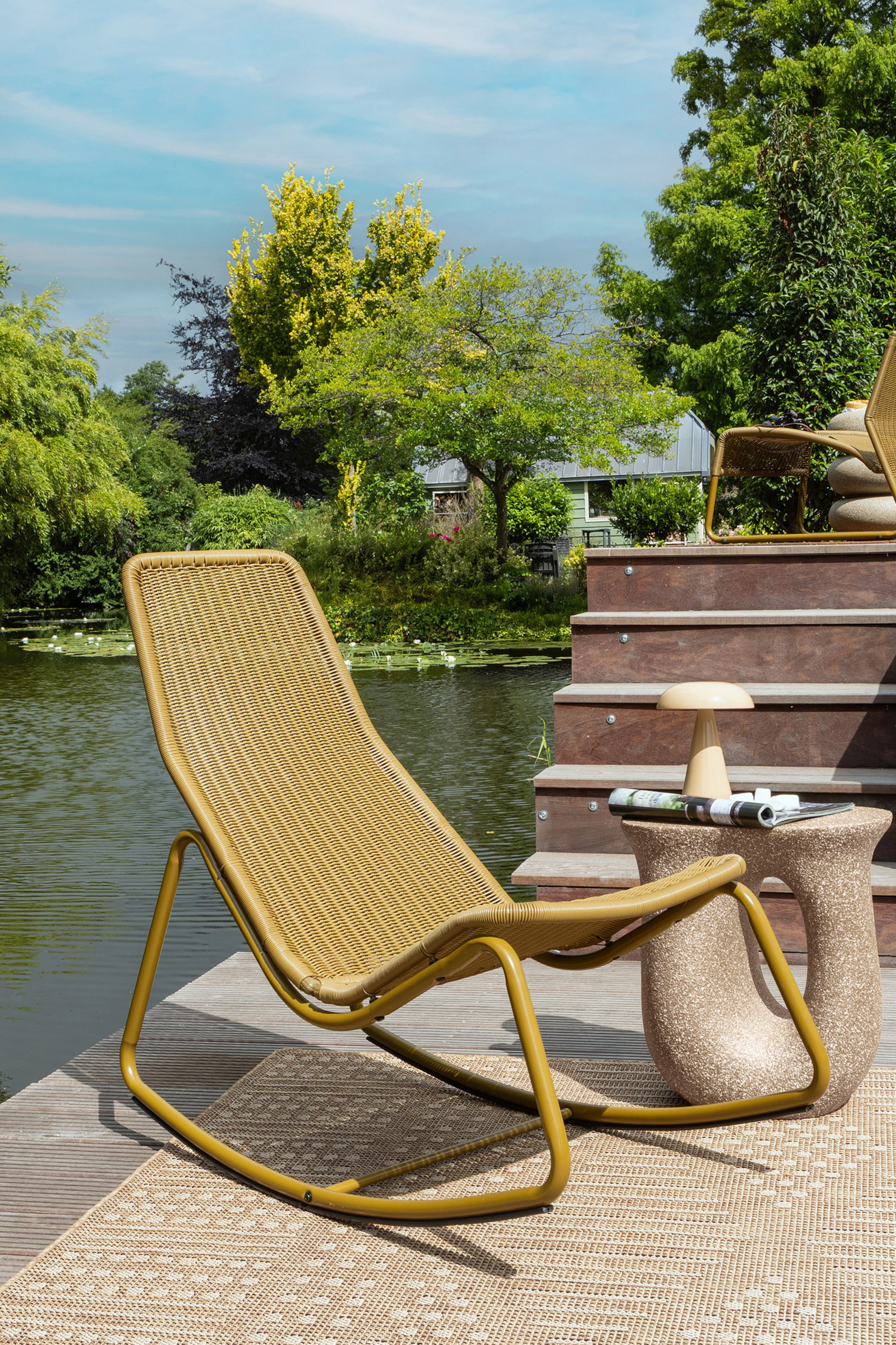 Agrandir l'image: Un fauteuil de salon jaune moutarde présente son profil complet, avec une surface d'assise tissée serrée et un cadre métallique courbé formant une base à bascule, reposant sur un tapis d'extérieur à motifs de couleur claire.